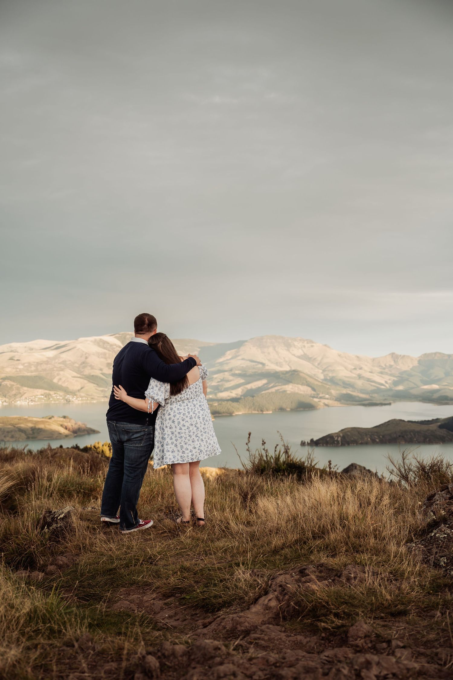 Couple embracing on hilltop overlooking scenic lake and snow-capped mountains under cloudy sky.