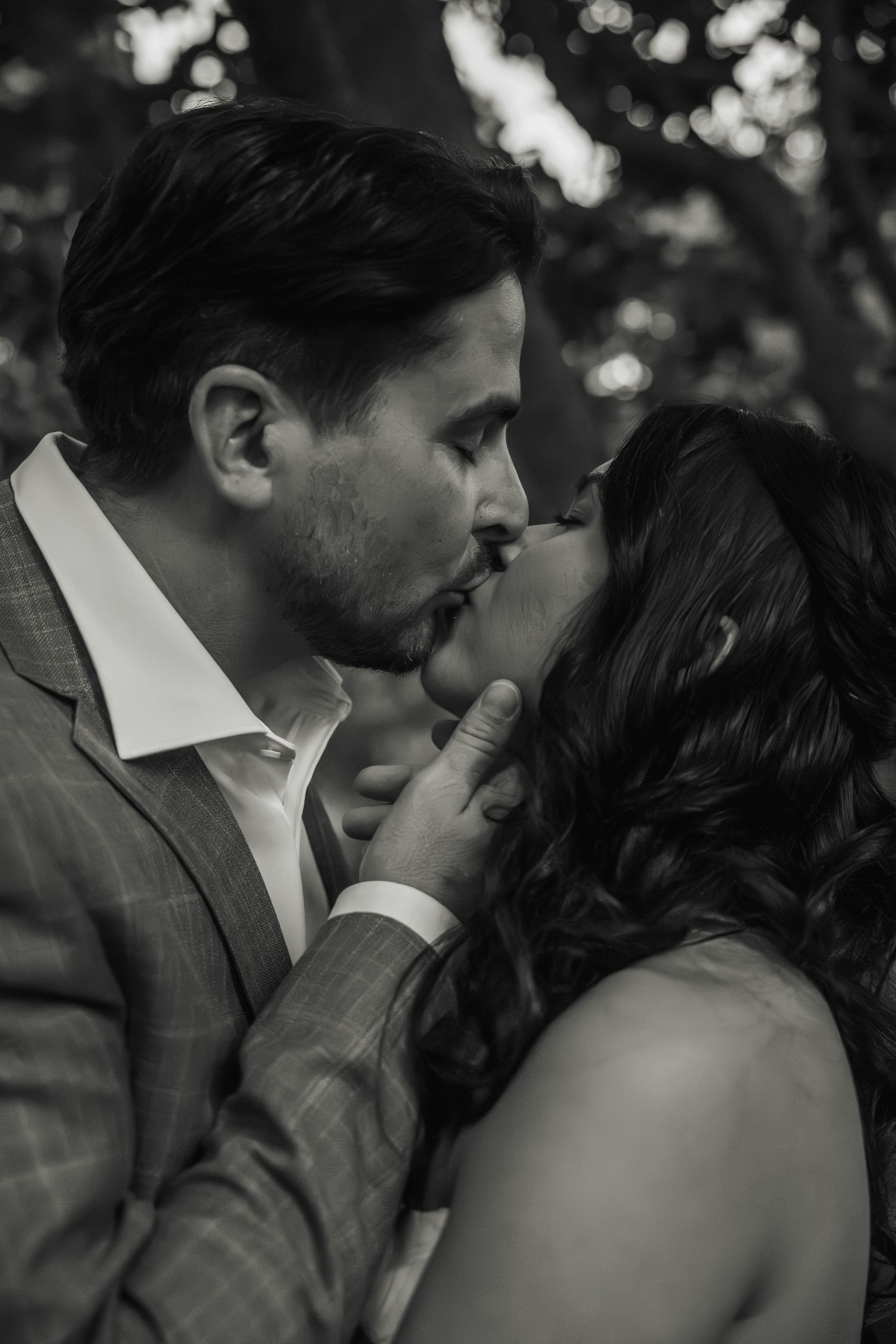 Black and white close-up of a bride and groom sharing a tender kiss outdoors among trees.