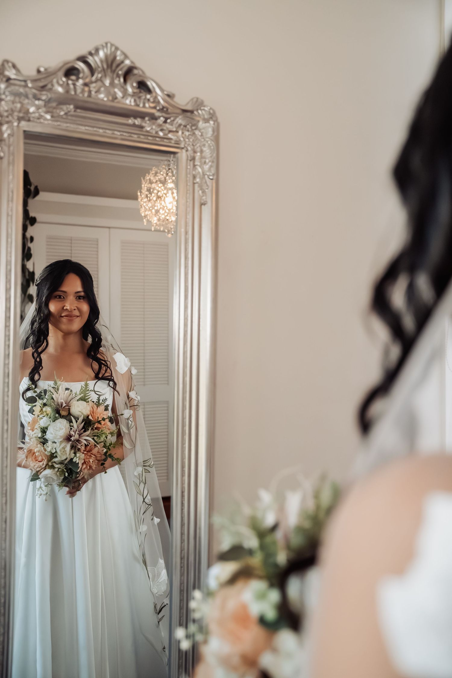 Bride in white gown holds floral bouquet while admiring her reflection in an ornate silver mirror.