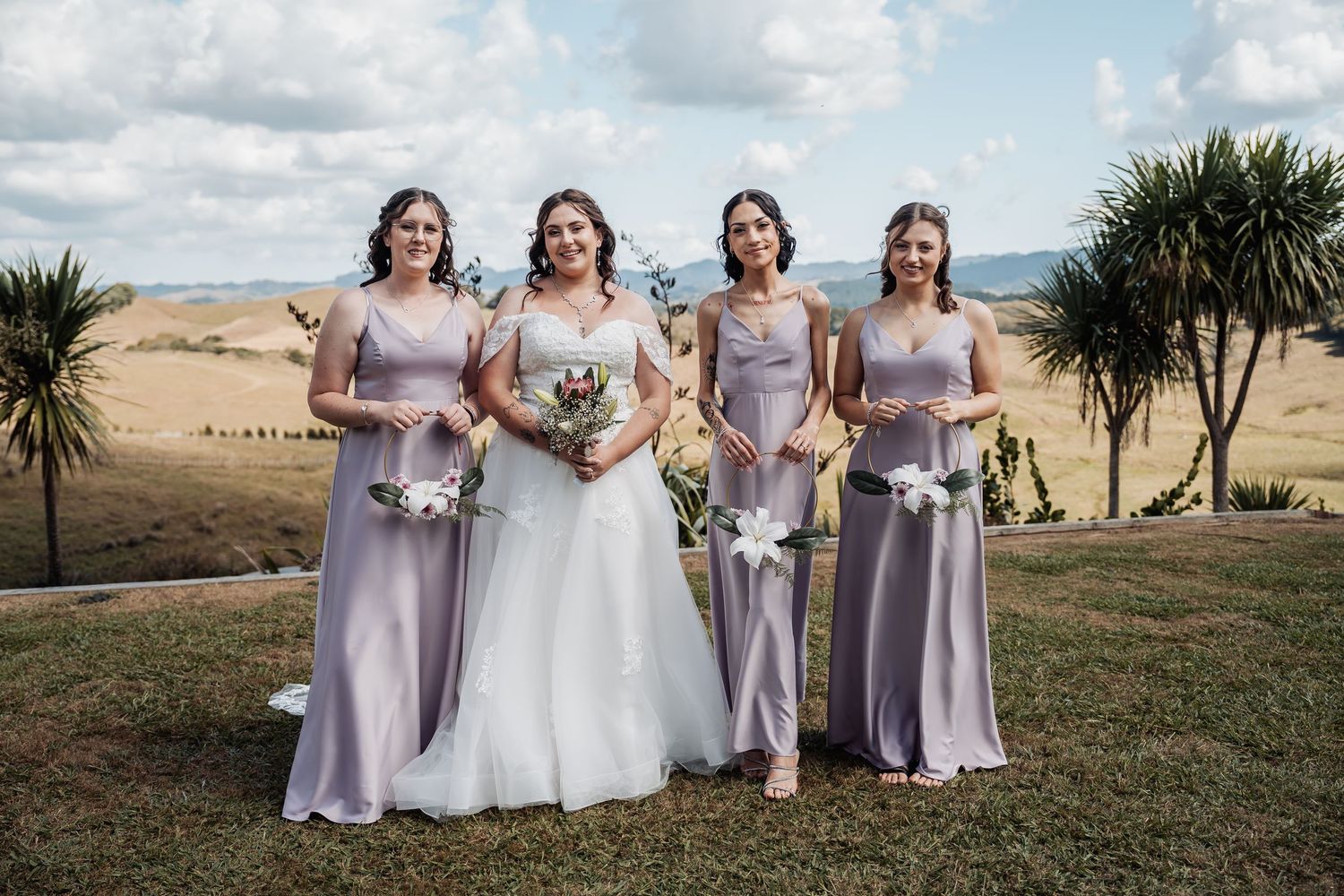 Bride in white gown with three bridesmaids in dusty purple dresses holding white bouquets outdoors.