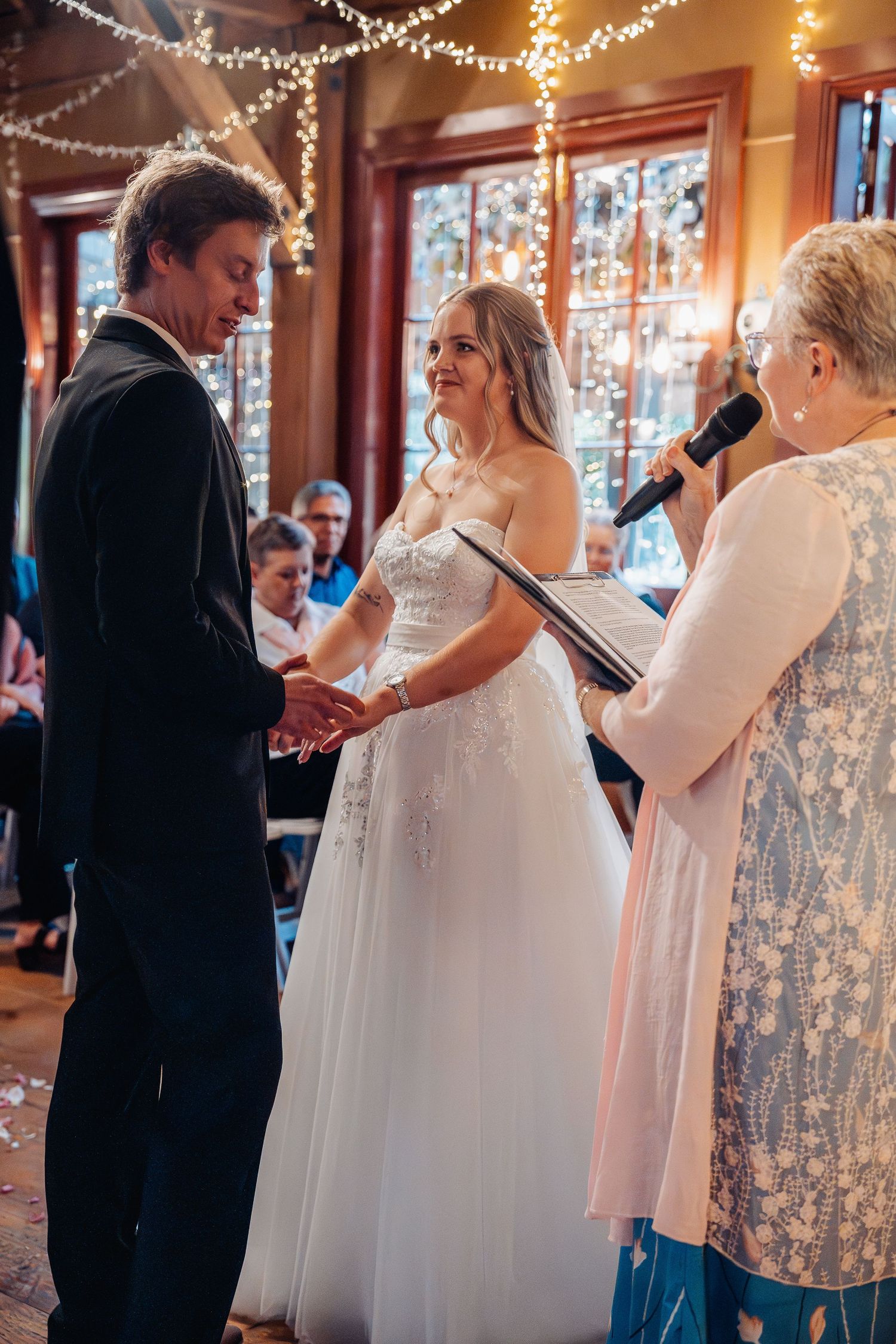 Wedding ceremony with couple exchanging vows in front of officiant, string lights and decor create warm glowing atmosphere.