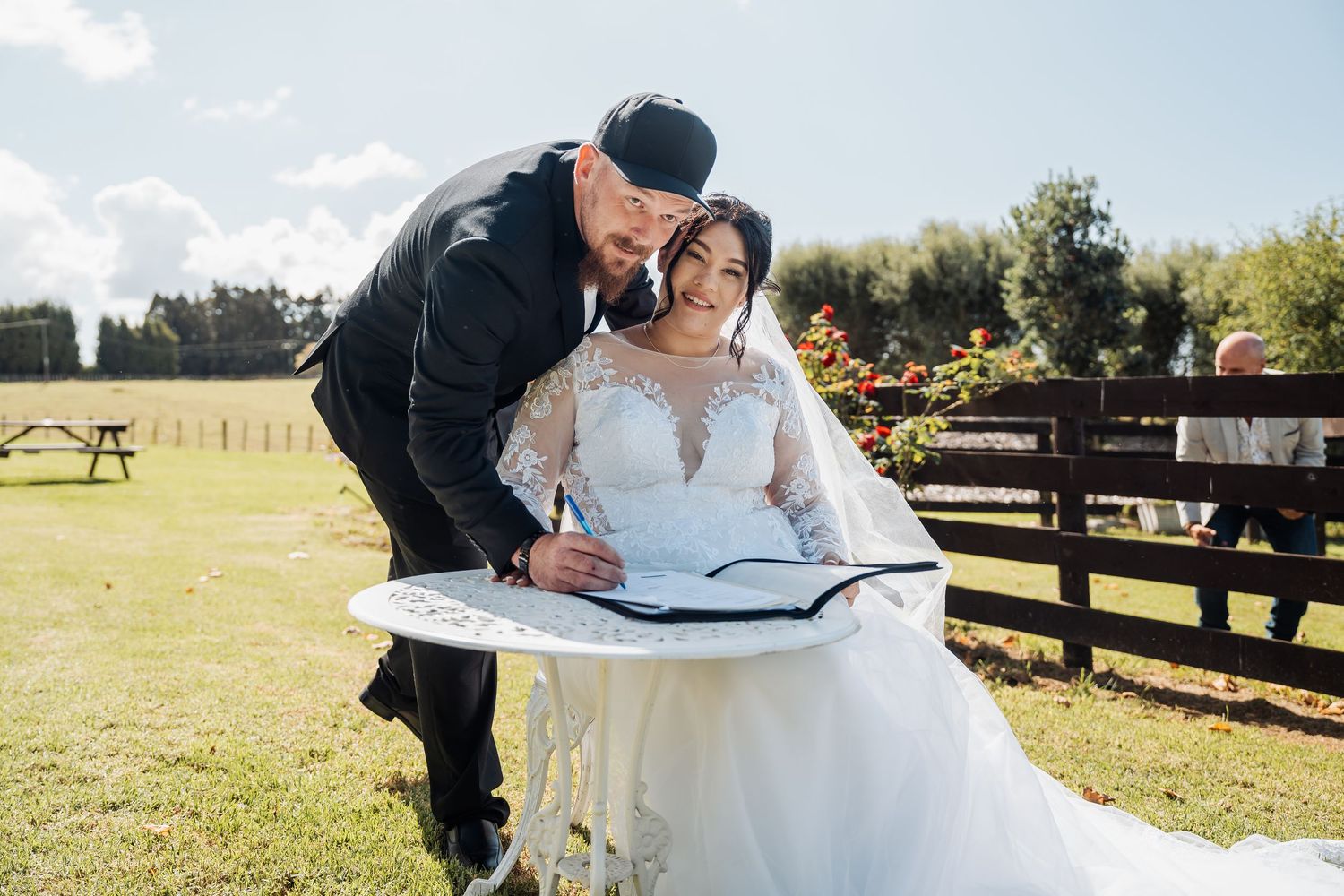 A couple signs their marriage certificate on a sunny outdoor wedding day with a farm fence in the background.