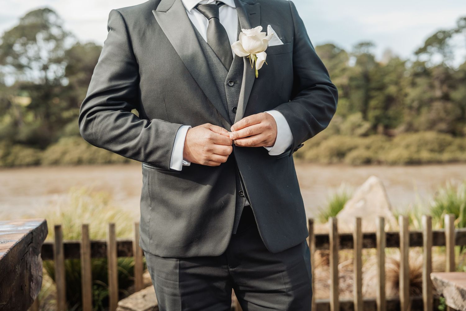 A single white boutonniere pin is shown being attached to a gray suit lapel at an outdoor wedding ceremony.