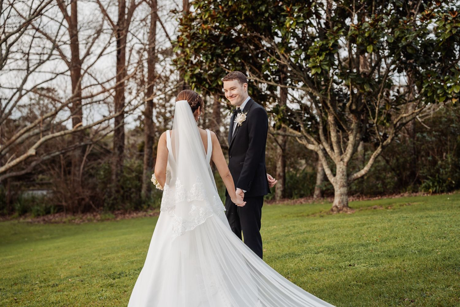 A white wedding gown billows gracefully in the breeze on a grassy lawn with trees.