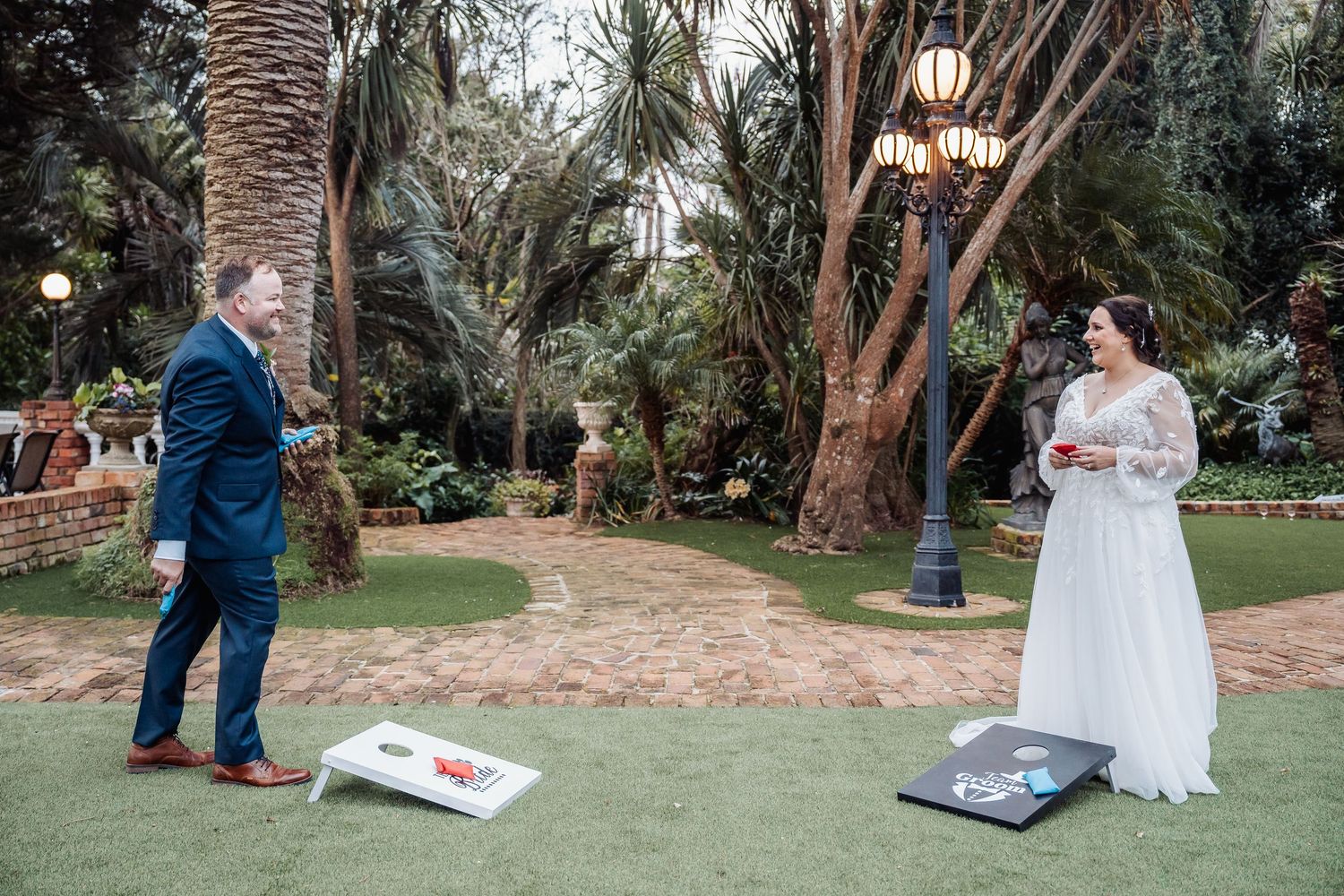 Wedding couple plays cornhole game outdoors surrounded by palm trees and string lights.