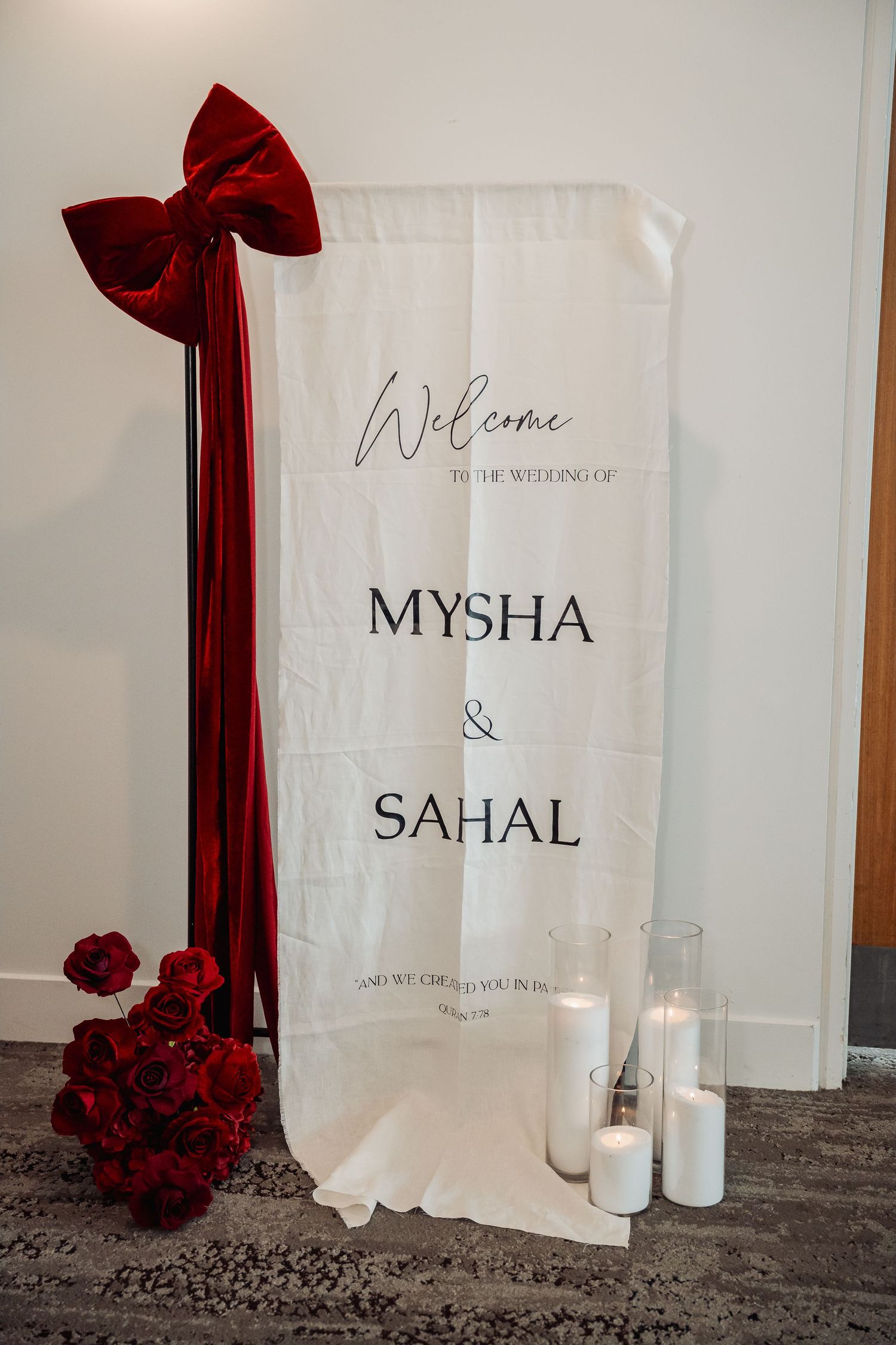 Wedding welcome sign with burgundy bow and red rose decorations against white backdrop.