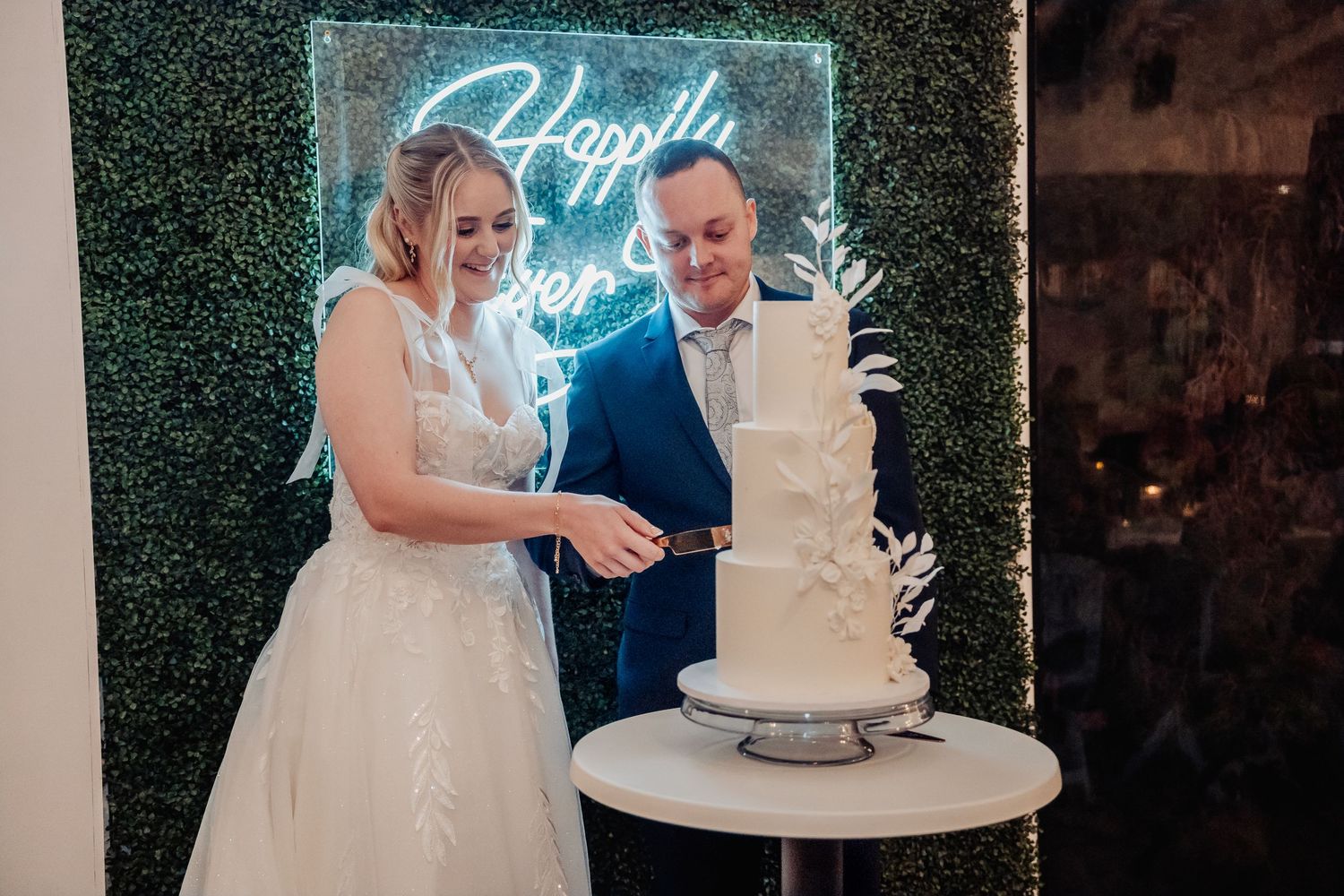 Wedding cake display with neon signage and greenery wall backdrop at reception.