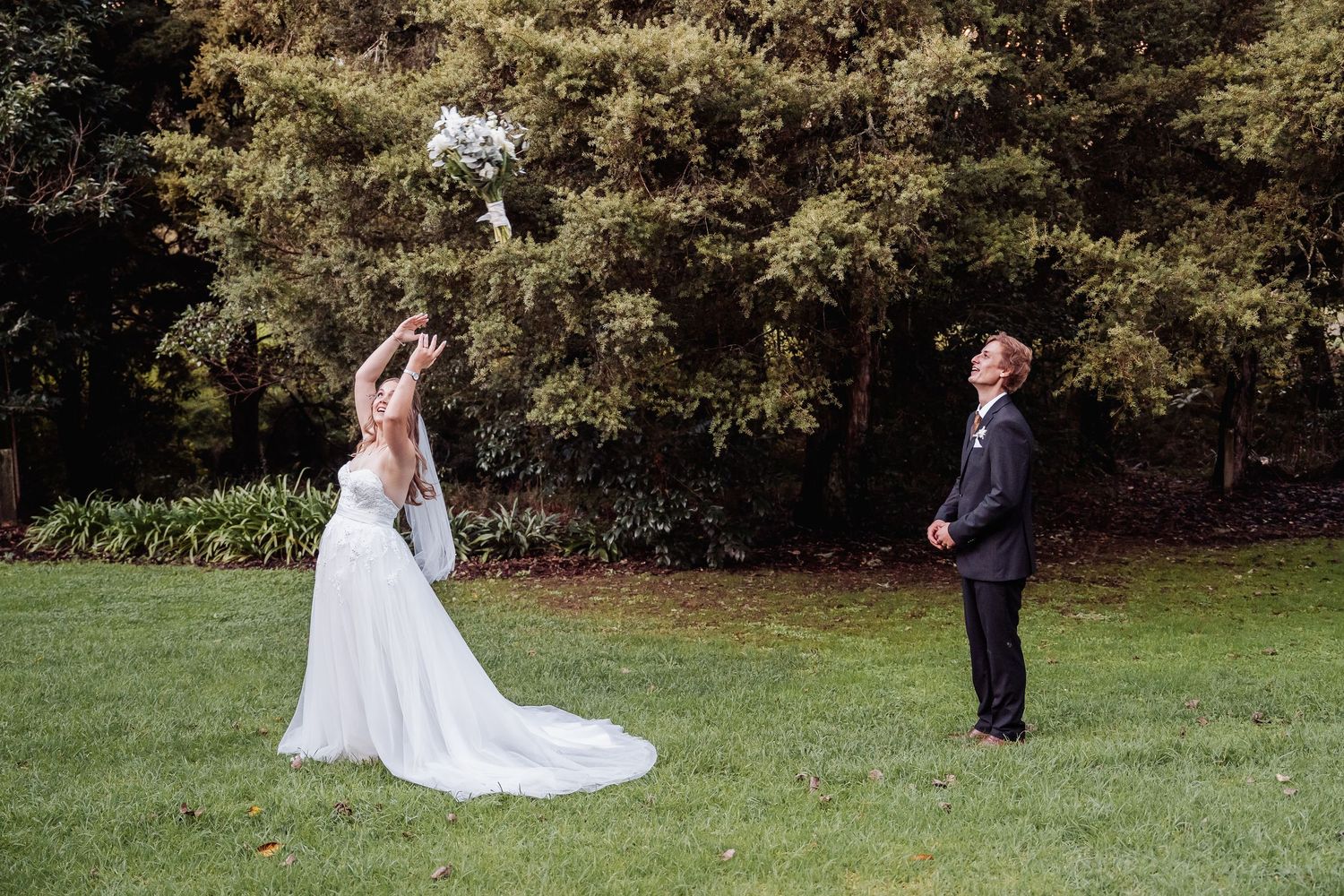 A couple plays a playful game of catch with their wedding bouquet on a grassy lawn surrounded by trees.