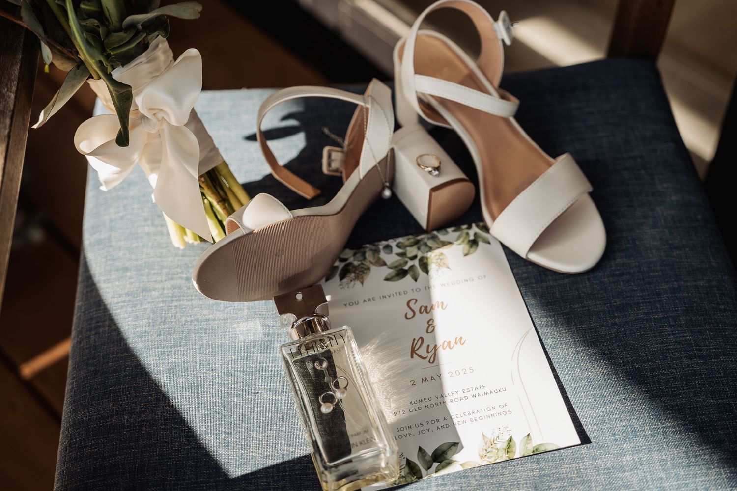 Wedding accessories including white sandals, perfume bottle and invitation card arranged on blue textured surface.