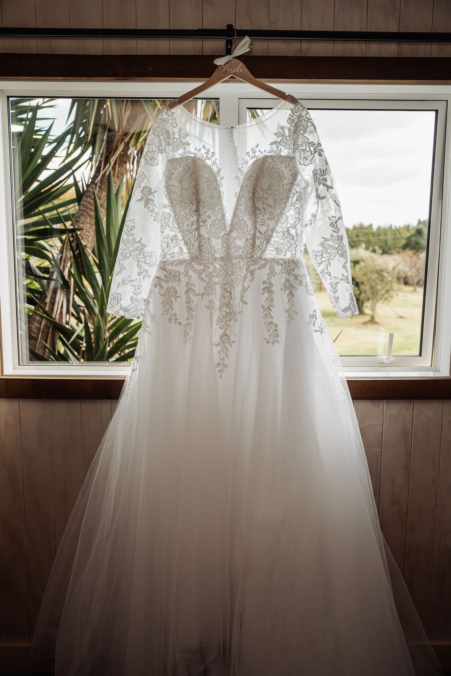 Elegant white wedding dress with lace details and long sleeves hanging by a window with palm leaves visible outside.