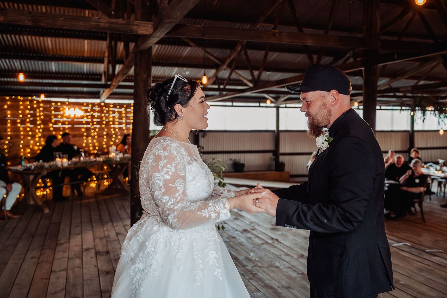 A couple shares their first dance during a rustic barn wedding reception with string lights and wooden decor.
