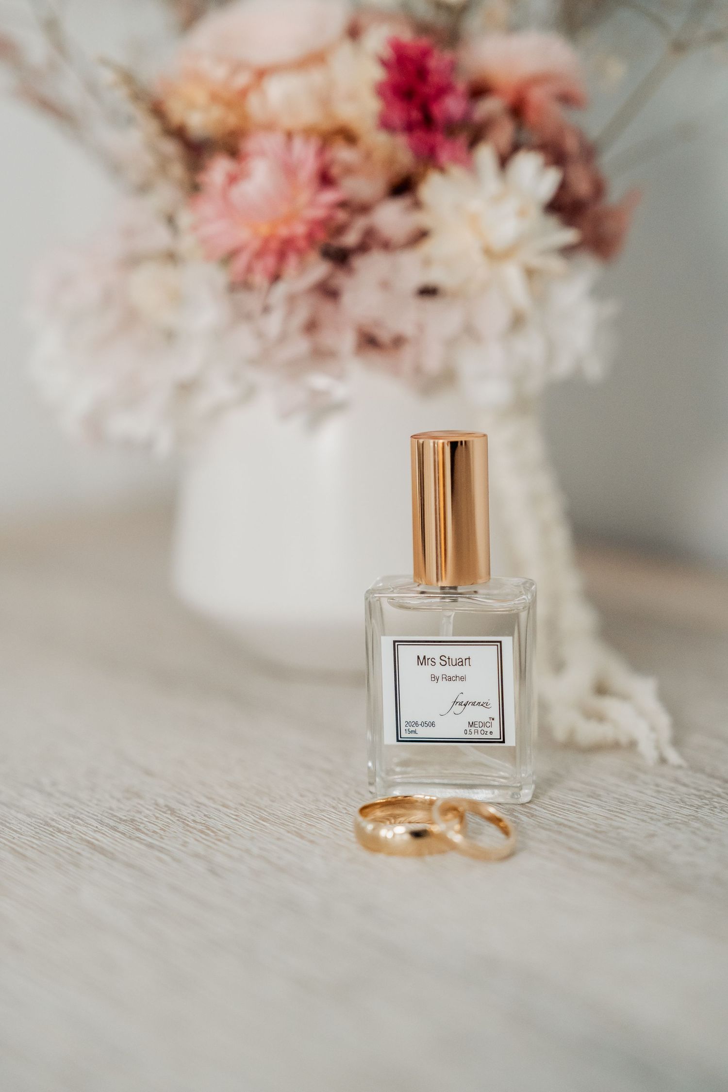 Wedding perfume bottle with gold rings and blush floral bouquet on white surface.