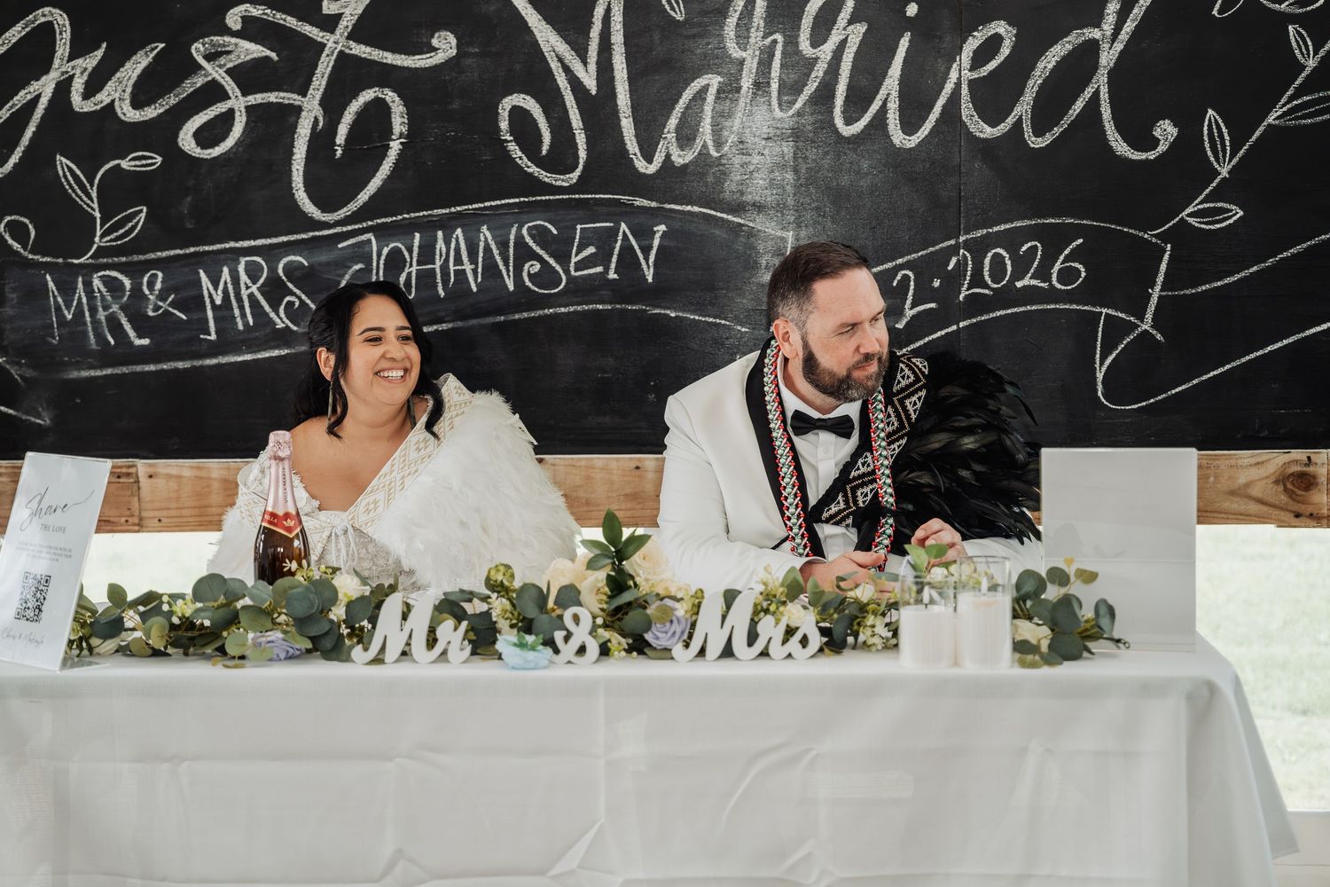 Bride and groom laugh at sweetheart table beneath Just Married Mr & Mrs Johansen chalkboard sign.