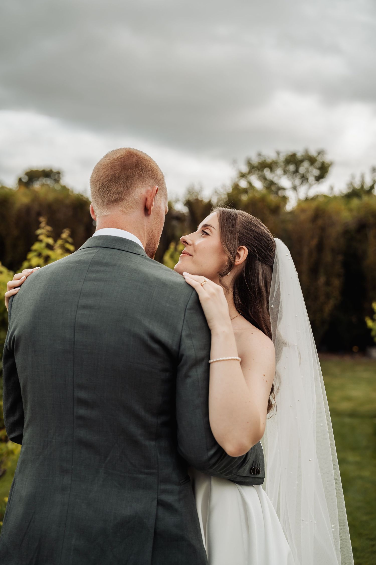 Bride in white gown and veil gazes lovingly at groom in gray suit against moody cloudy backdrop.