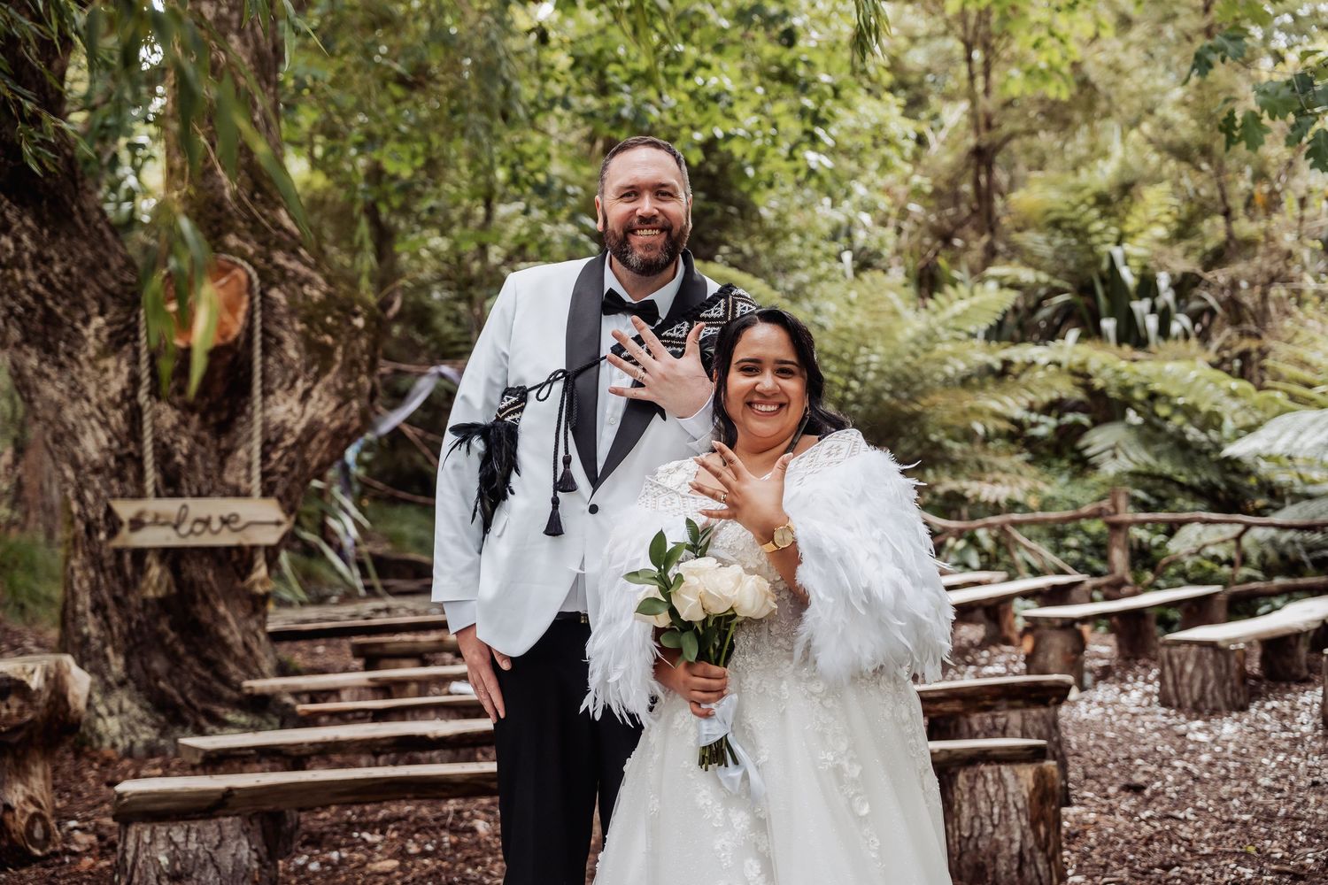 Smiling bride and groom pose together in lush forest setting, bride holding white bouquet.