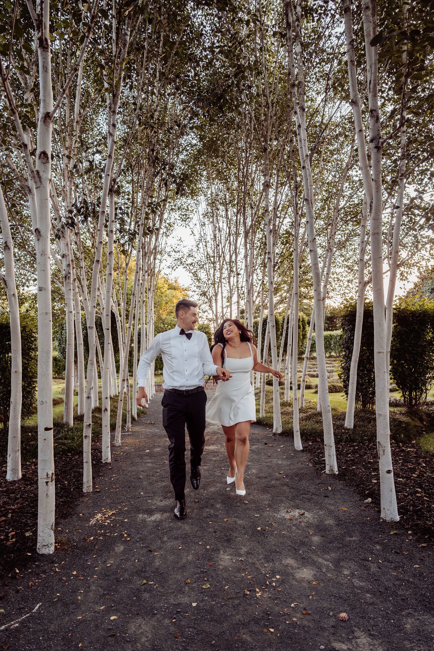 Bride and groom holding hands and walking joyfully through an elegant row of white birch trees.