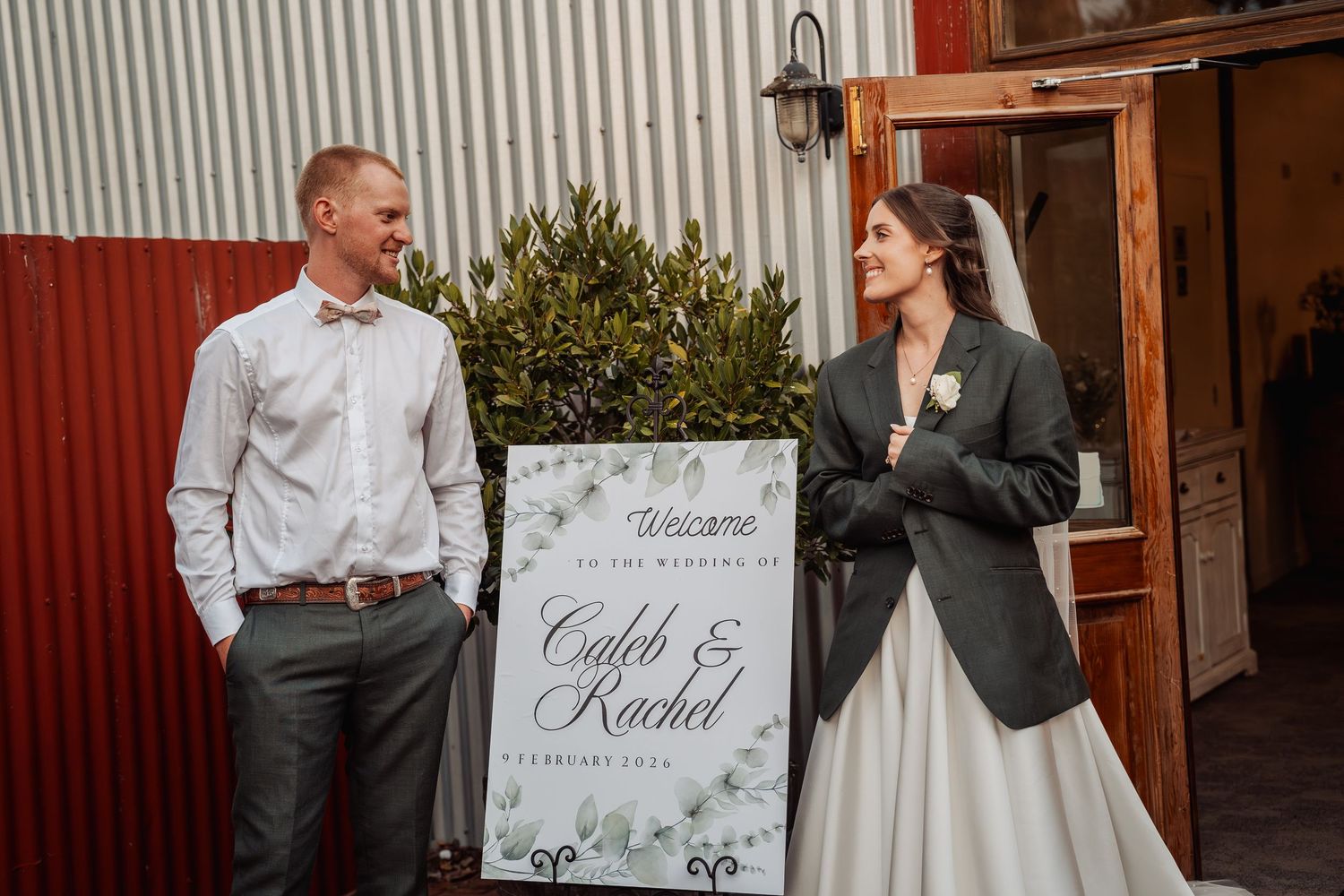 Bride and groom standing beside a greenery wedding welcome sign reading Caleb and Rachel.