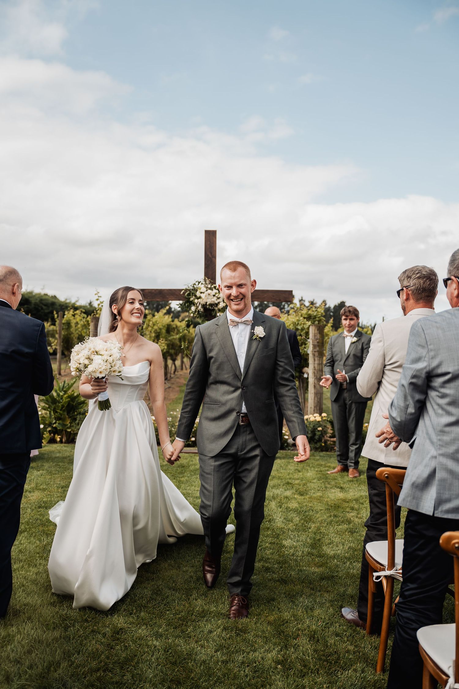 Joyful bride and groom walking down the aisle after outdoor vineyard wedding ceremony under a blue sky.