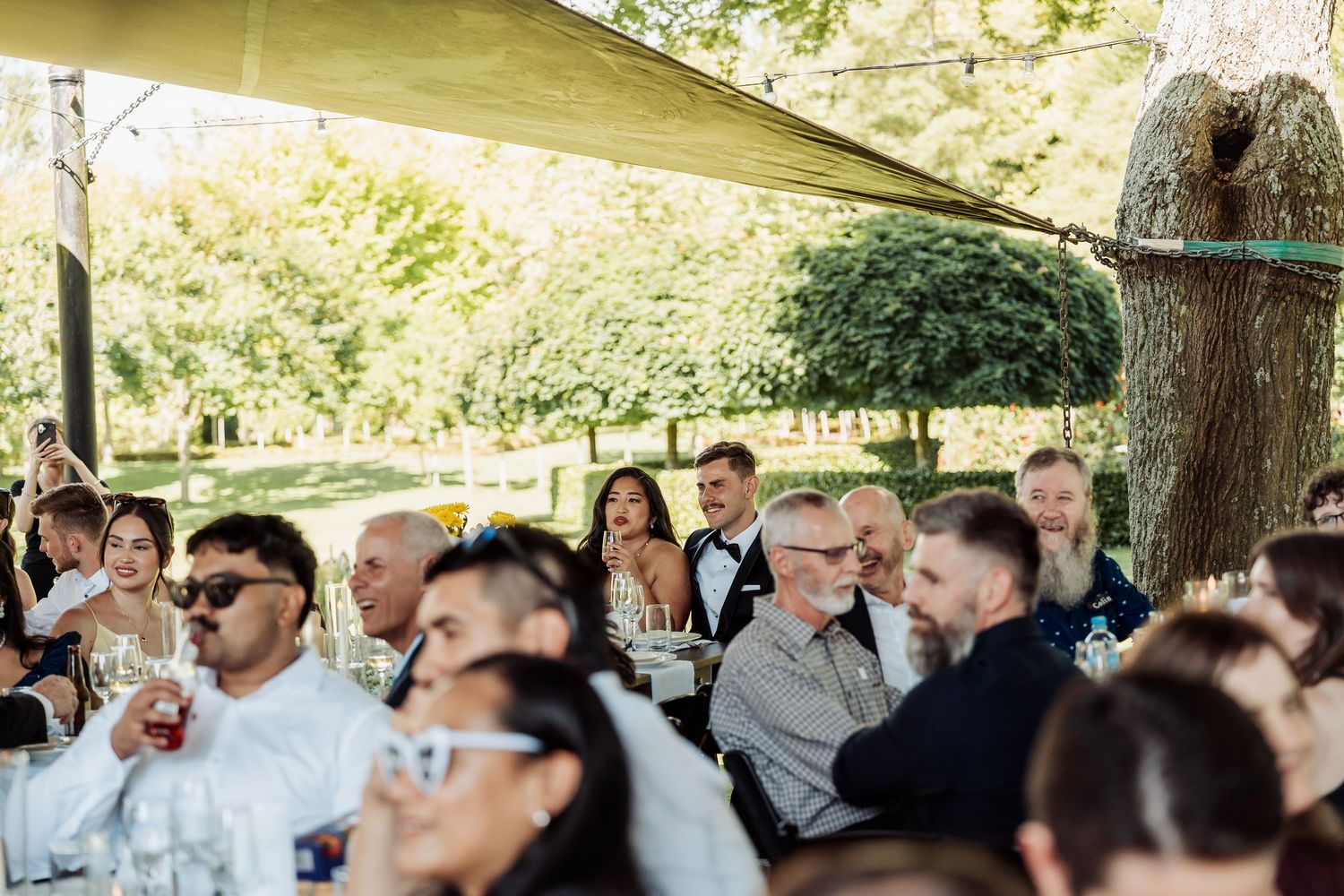 Wedding guests seated outdoors under canopy shade enjoying reception celebration surrounded by lush green garden trees.
