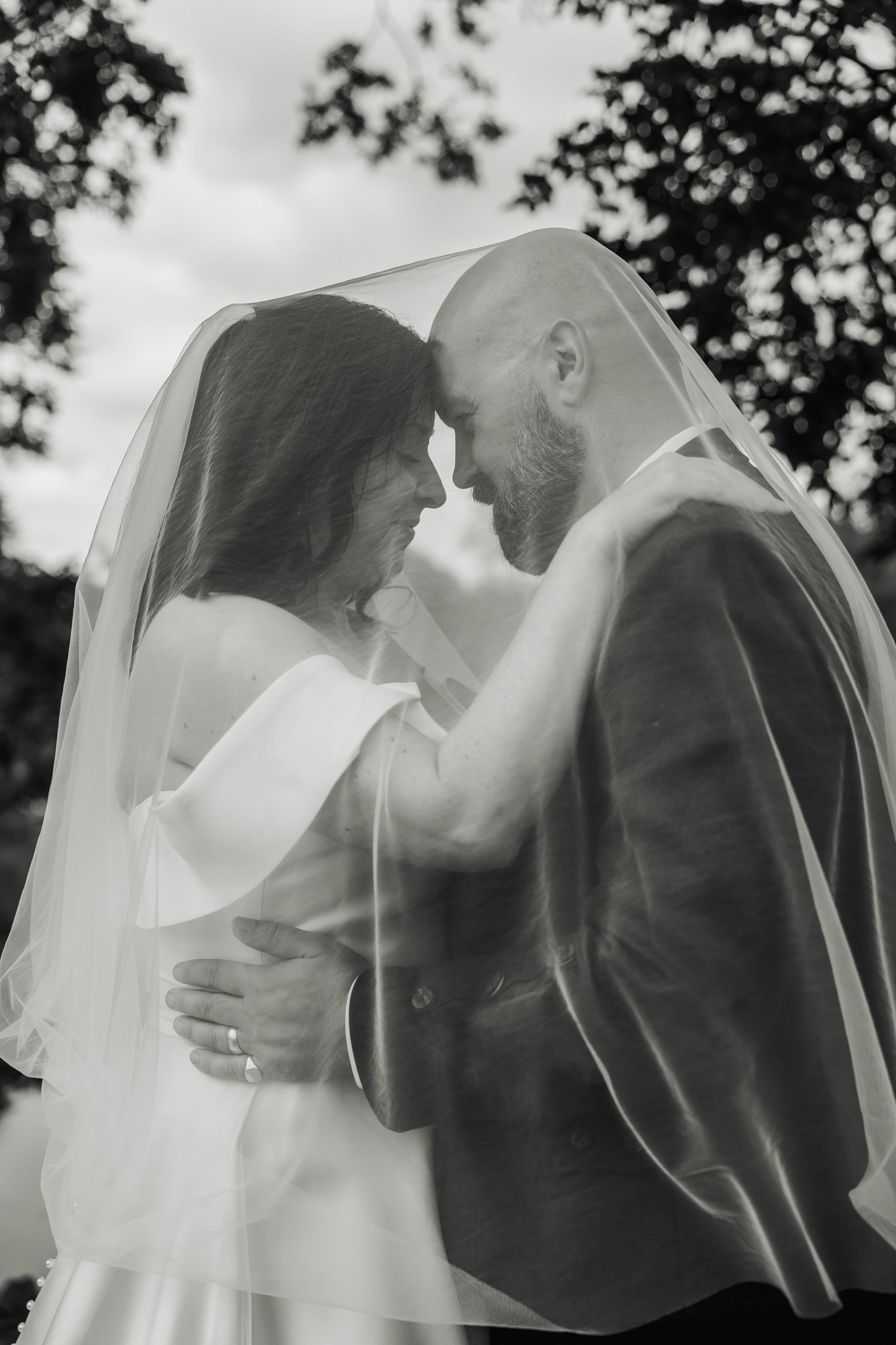 Black and white romantic portrait of bride and groom embracing tenderly beneath flowing wedding veil outdoors.