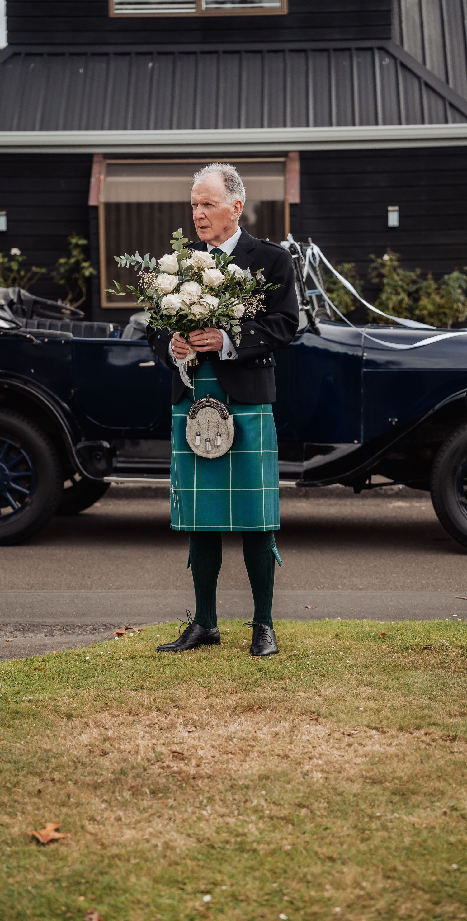 Man in green tartan kilt holds white rose bouquet beside a vintage black wedding car.