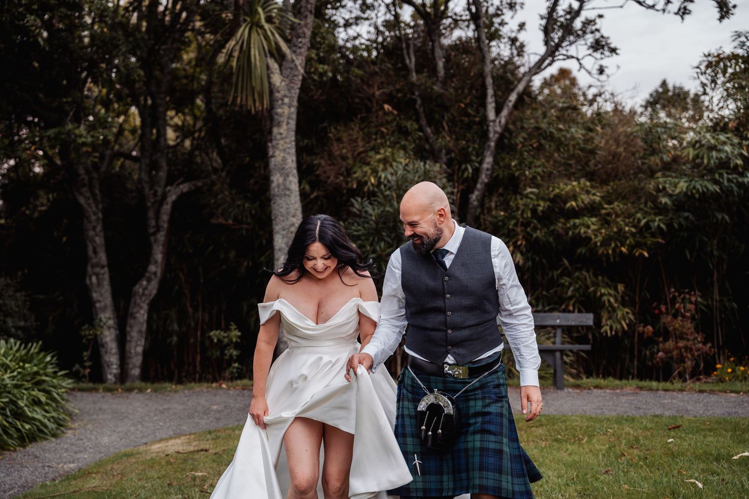 Bride lifting her white gown while walking with her kilted groom through a green garden path.