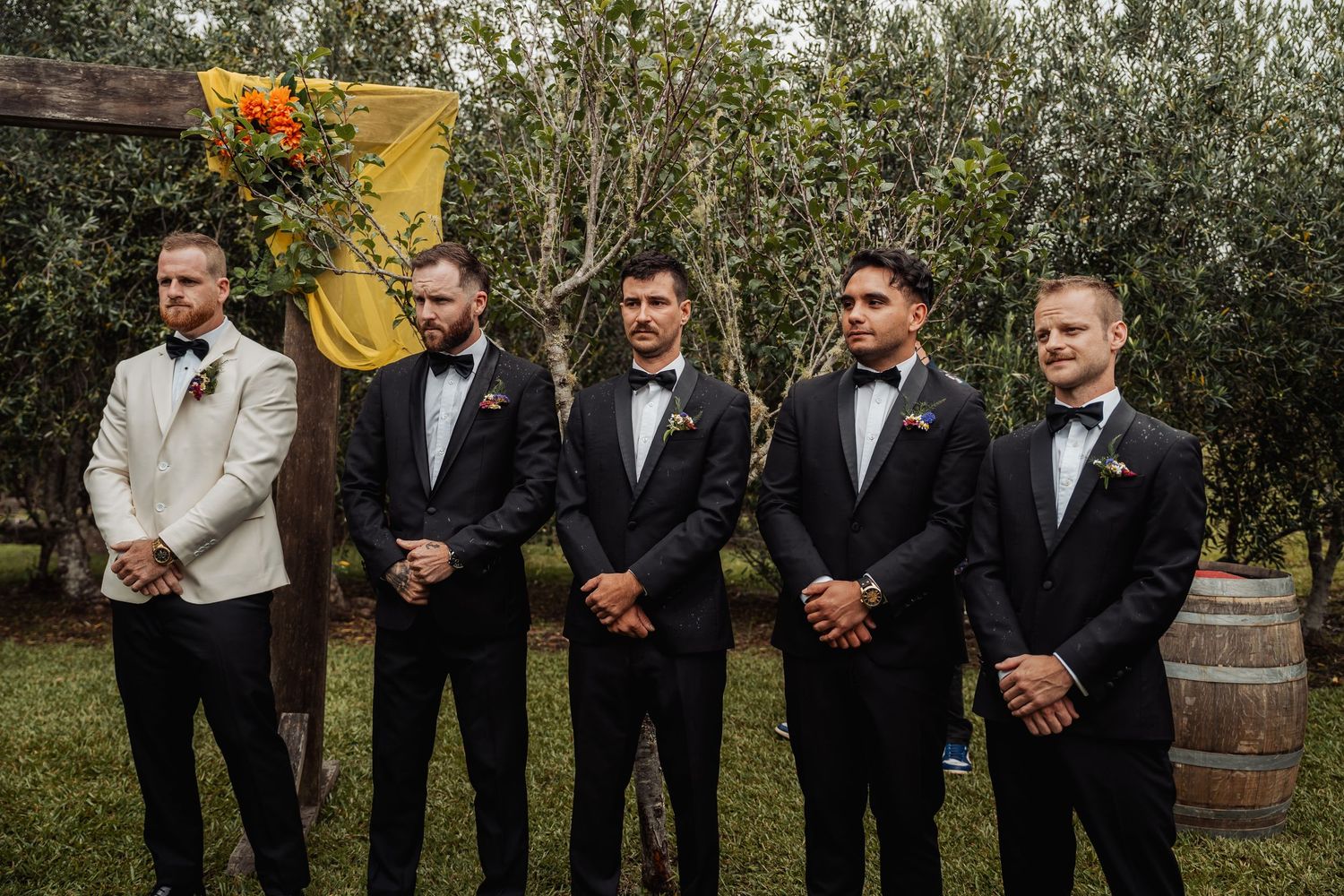 Five groomsmen in black tuxedos with bow ties standing at an outdoor wedding ceremony.