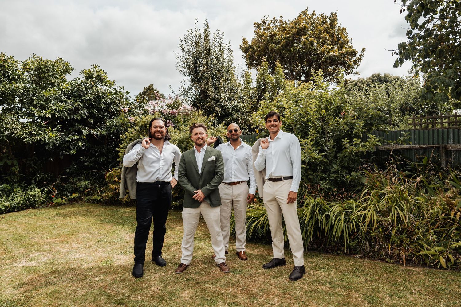 Four groomsmen in suits pose together smiling in a lush garden before a wedding ceremony.