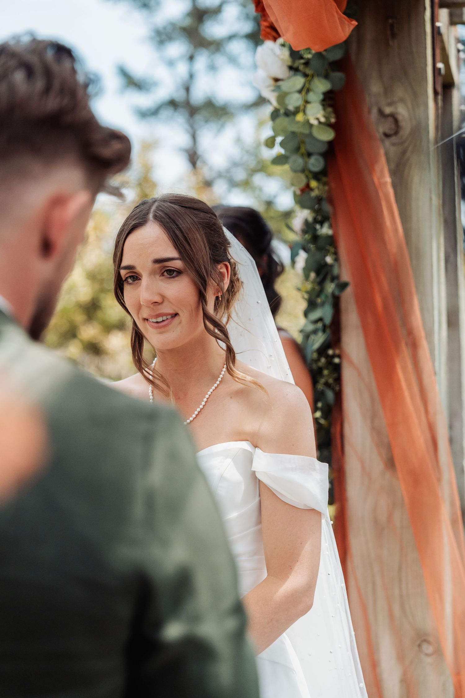 Emotional bride in off-shoulder white gown gazes at groom during outdoor wedding ceremony.