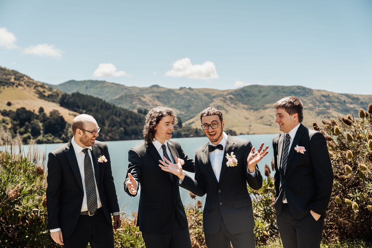 Groom and groomsmen in black suits laughing outdoors with scenic lake and mountains in New Zealand.