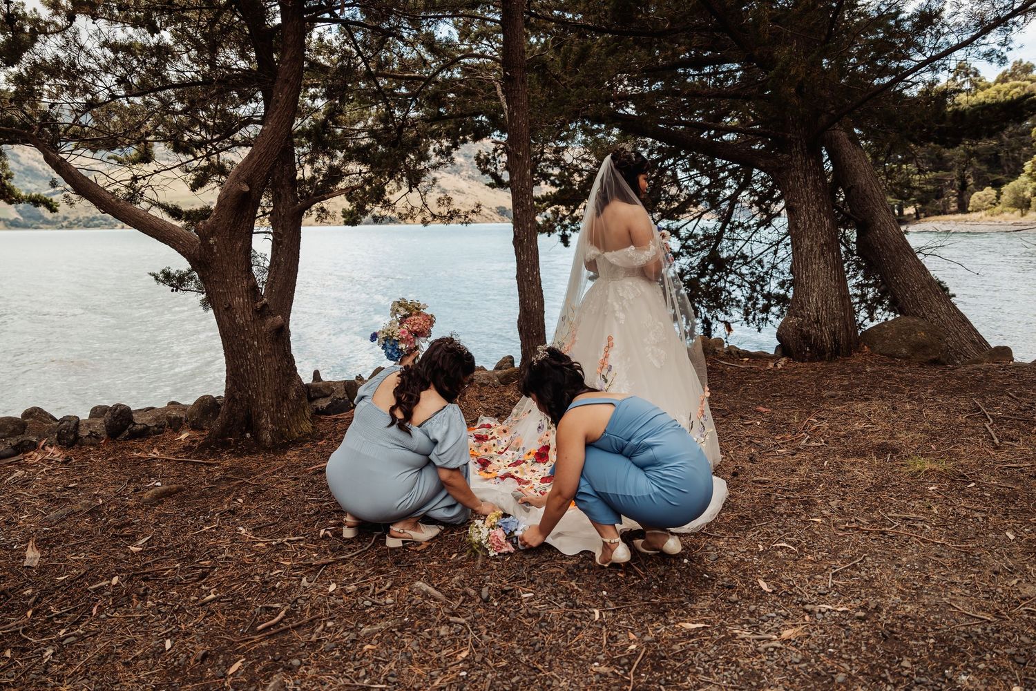 Bridesmaids helping bride with dress adjustments under pine trees near a scenic blue lake.