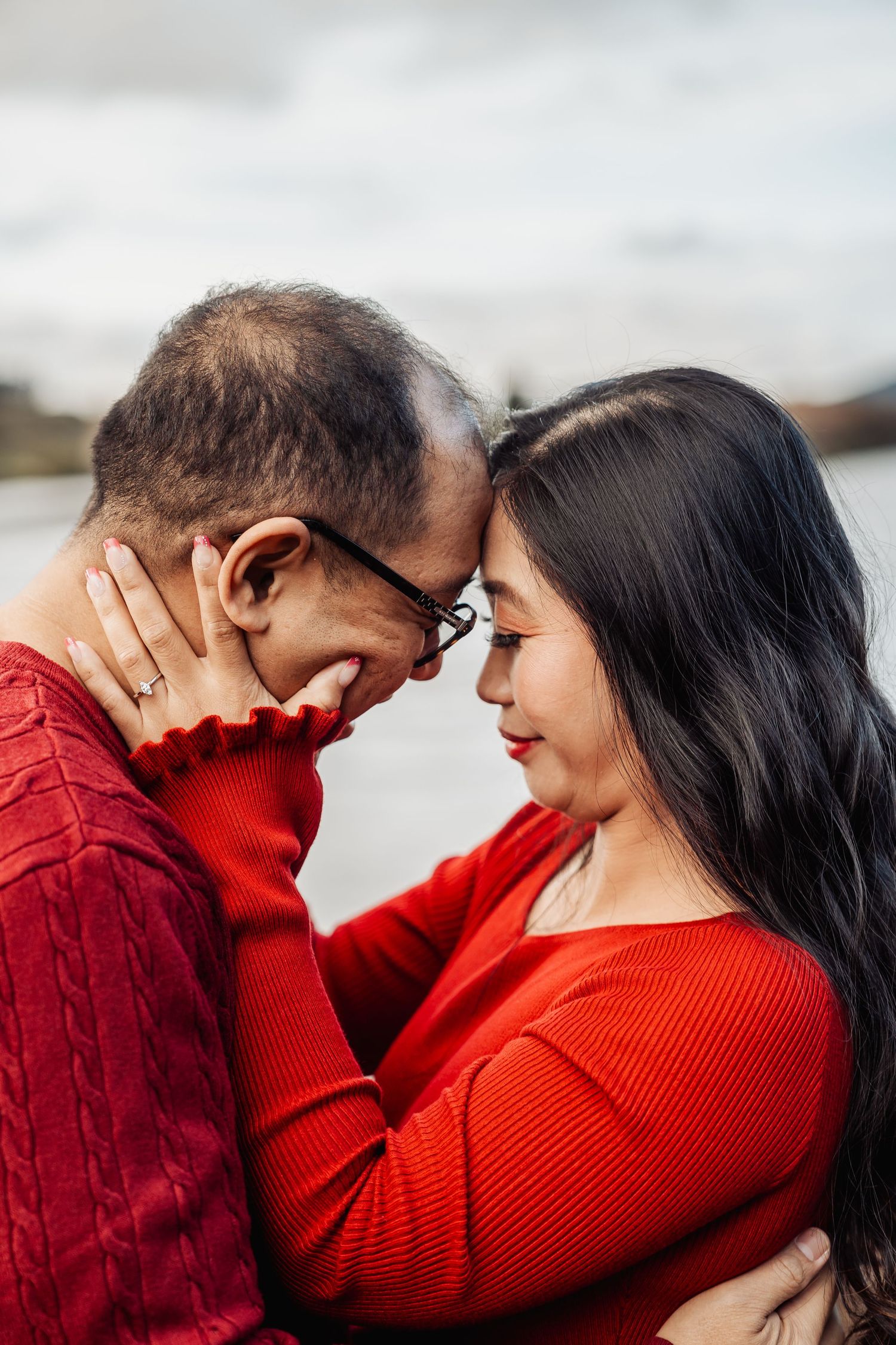 Couple in matching red sweaters share an intimate forehead touch outdoors near a lake.