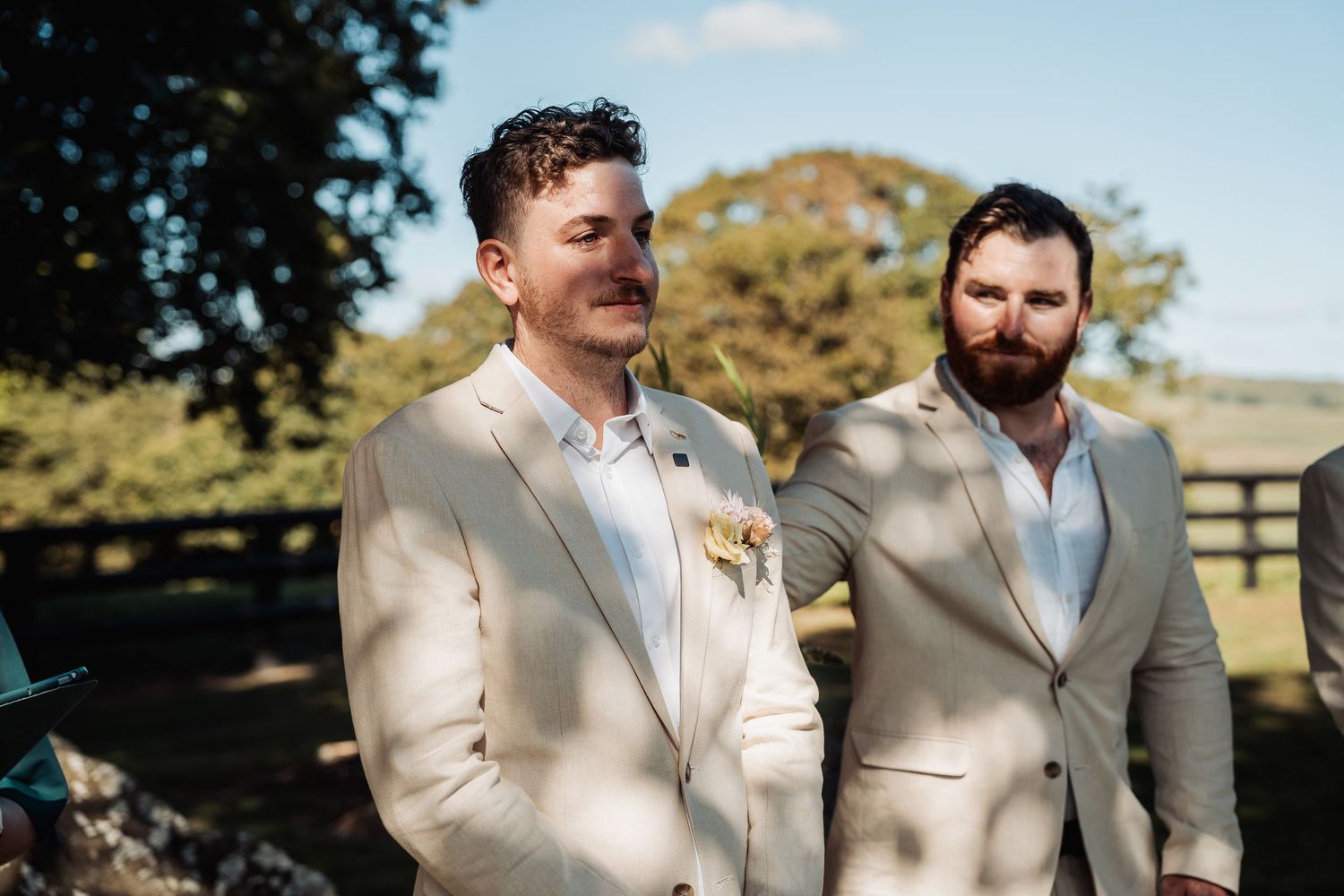 Groom and best man in beige suits waiting at outdoor wedding ceremony altar.