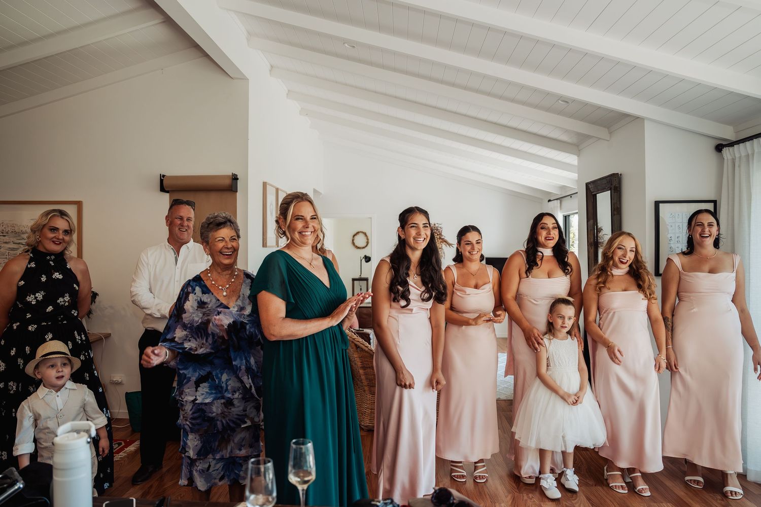 Bride in white gown with bridesmaids in blush pink dresses and family gathered inside a wedding venue.