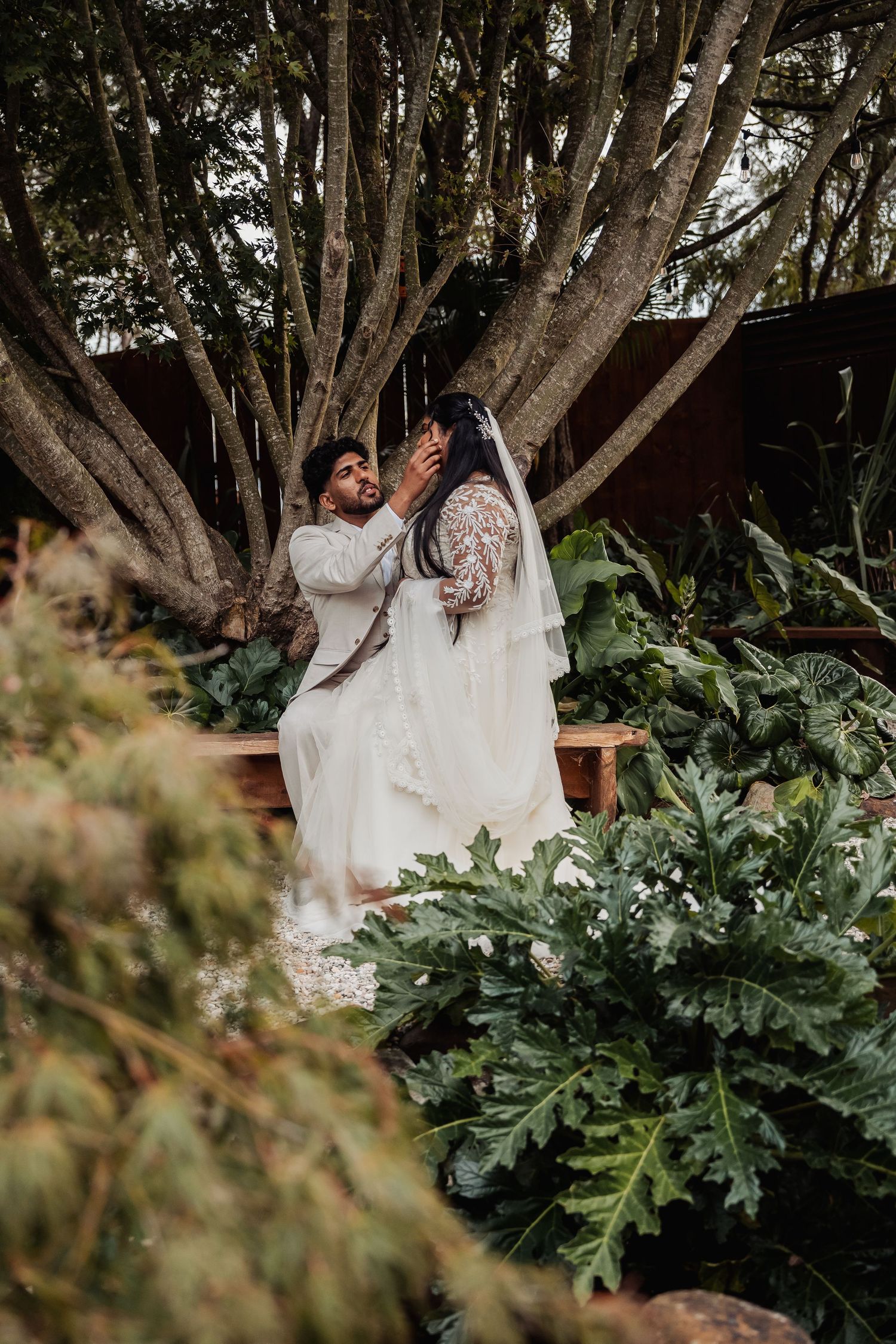 Groom tenderly touches brides veil among lush garden greenery beside a large tree.