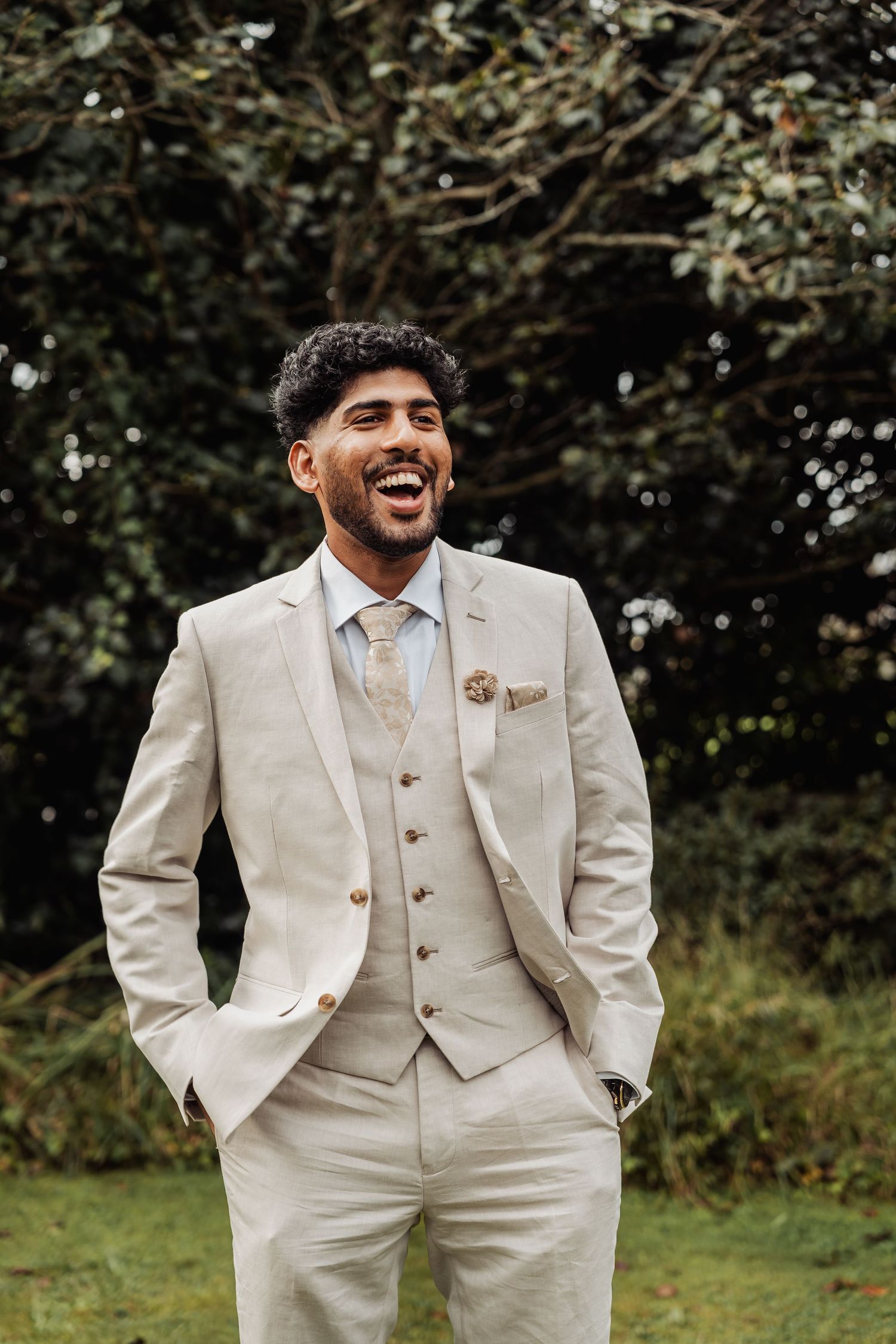 Smiling groom in beige three-piece suit with floral lapel pin standing outdoors at wedding.