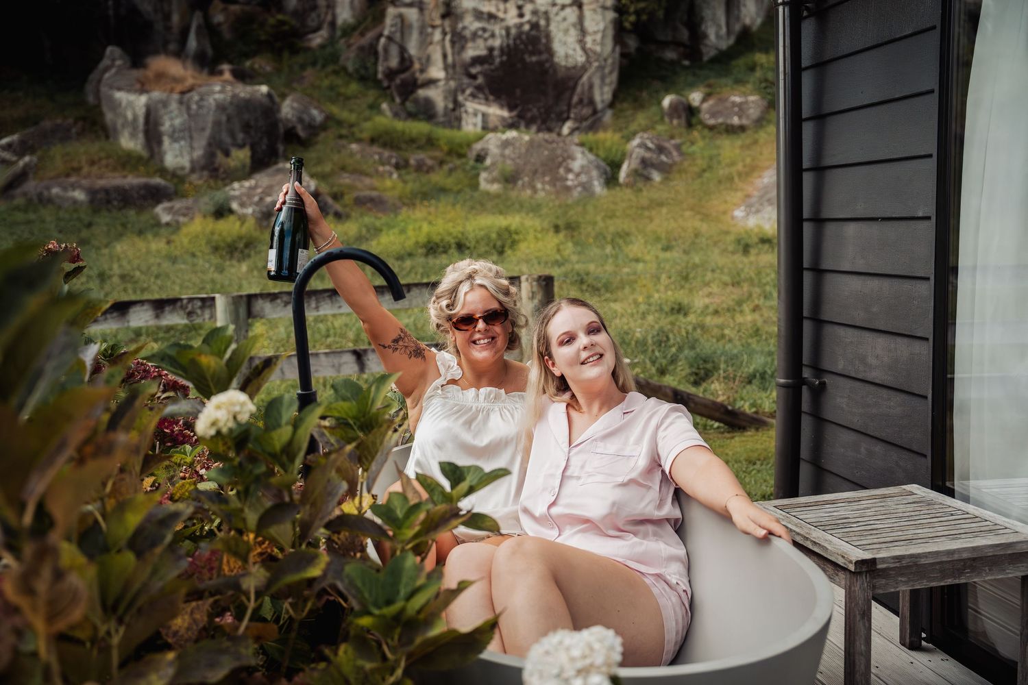 Two women smiling and relaxing in an outdoor bathtub surrounded by lush garden greenery.