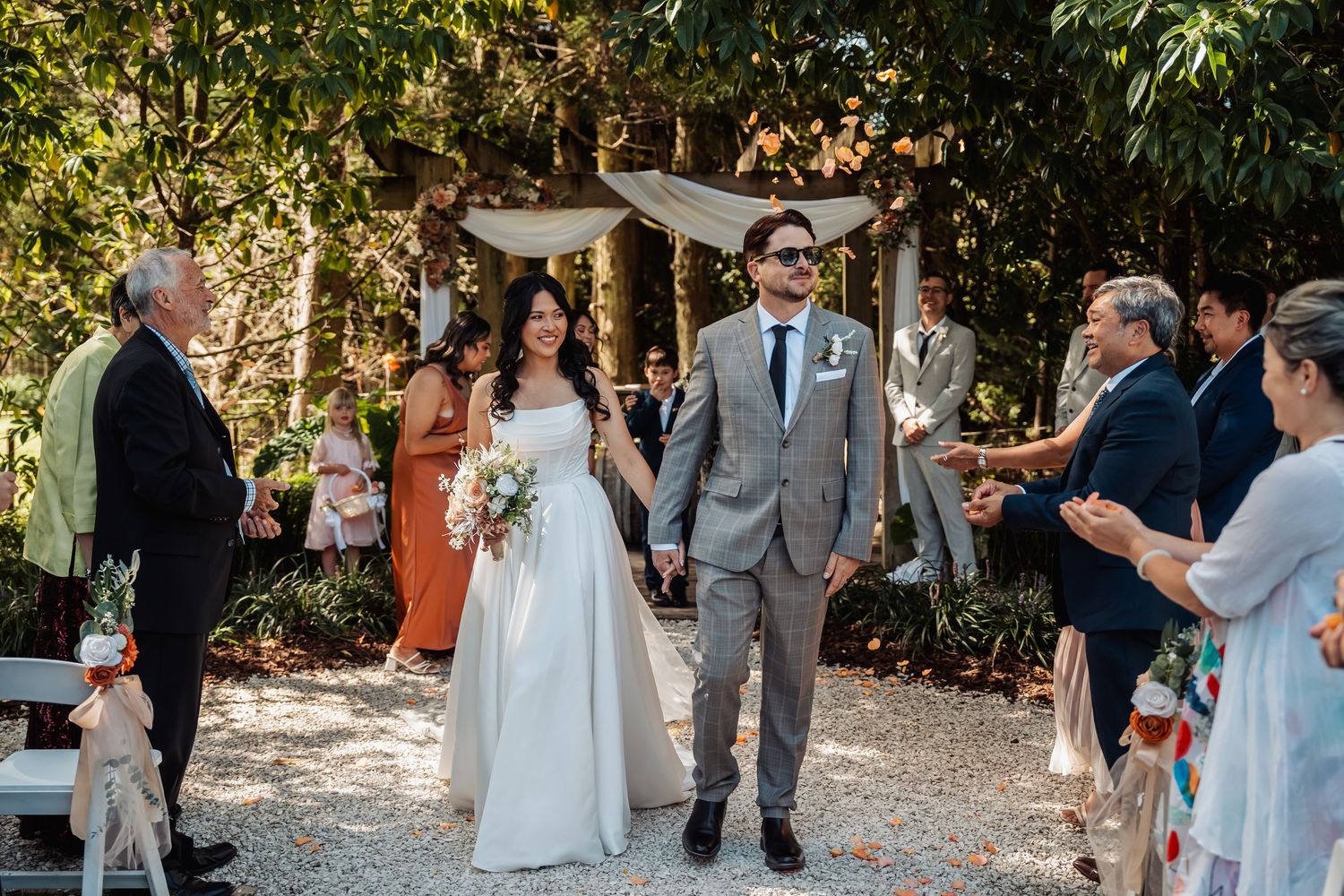 Newlyweds walk back down aisle hand in hand as guests celebrate with sparklers in sunny garden.