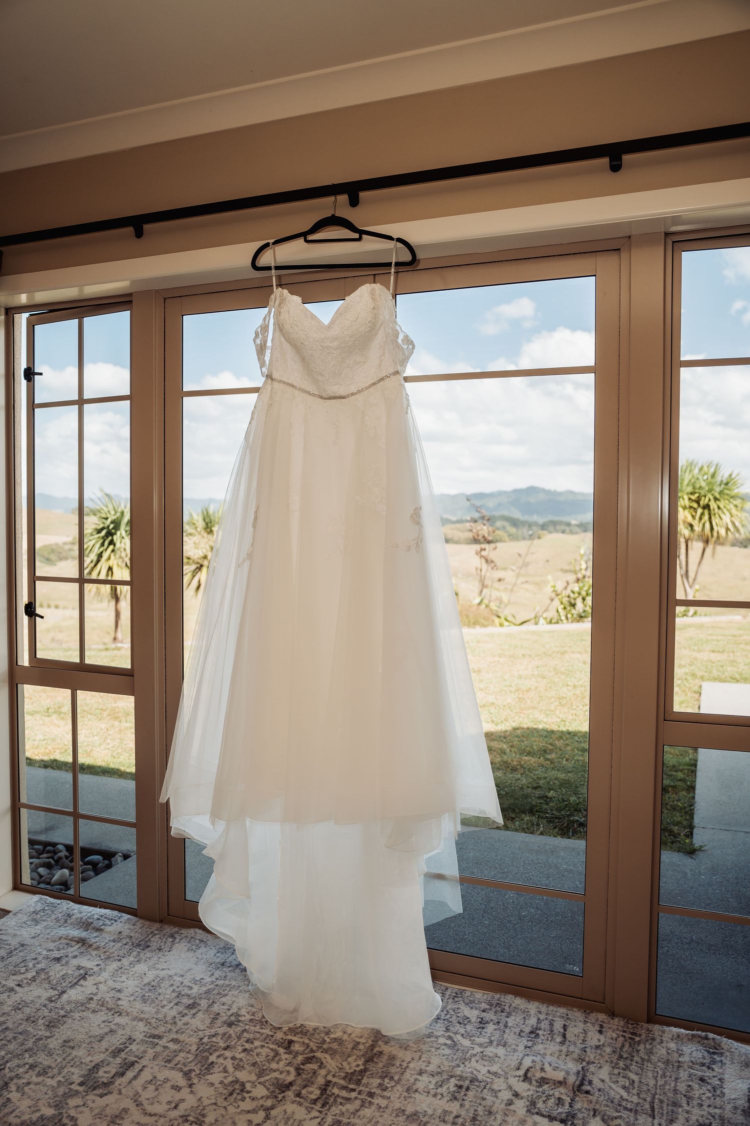 White wedding gown hanging on a hanger in front of French doors with scenic countryside views.