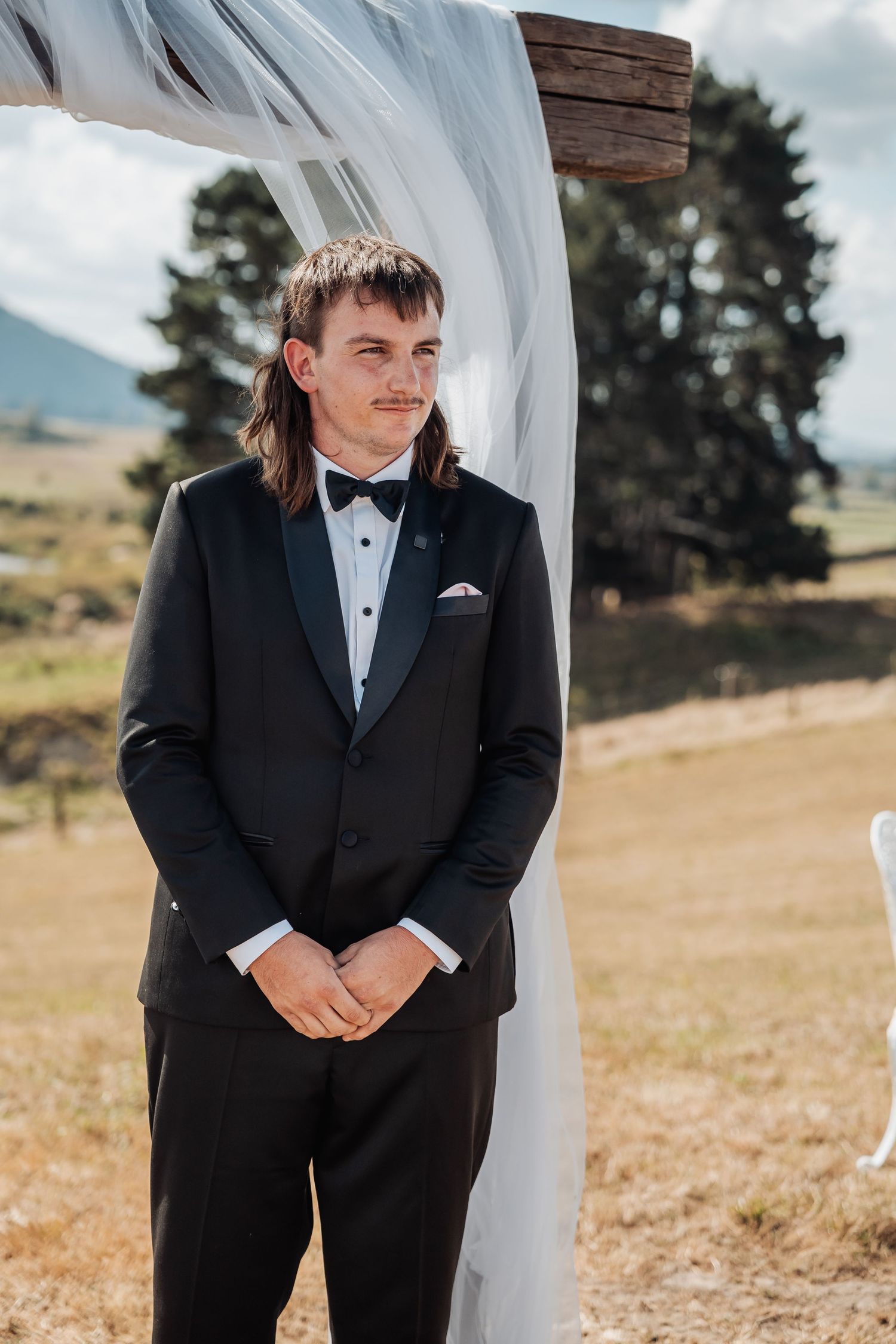 Groom in black tuxedo with bow tie standing at outdoor wedding altar with scenic countryside backdrop.