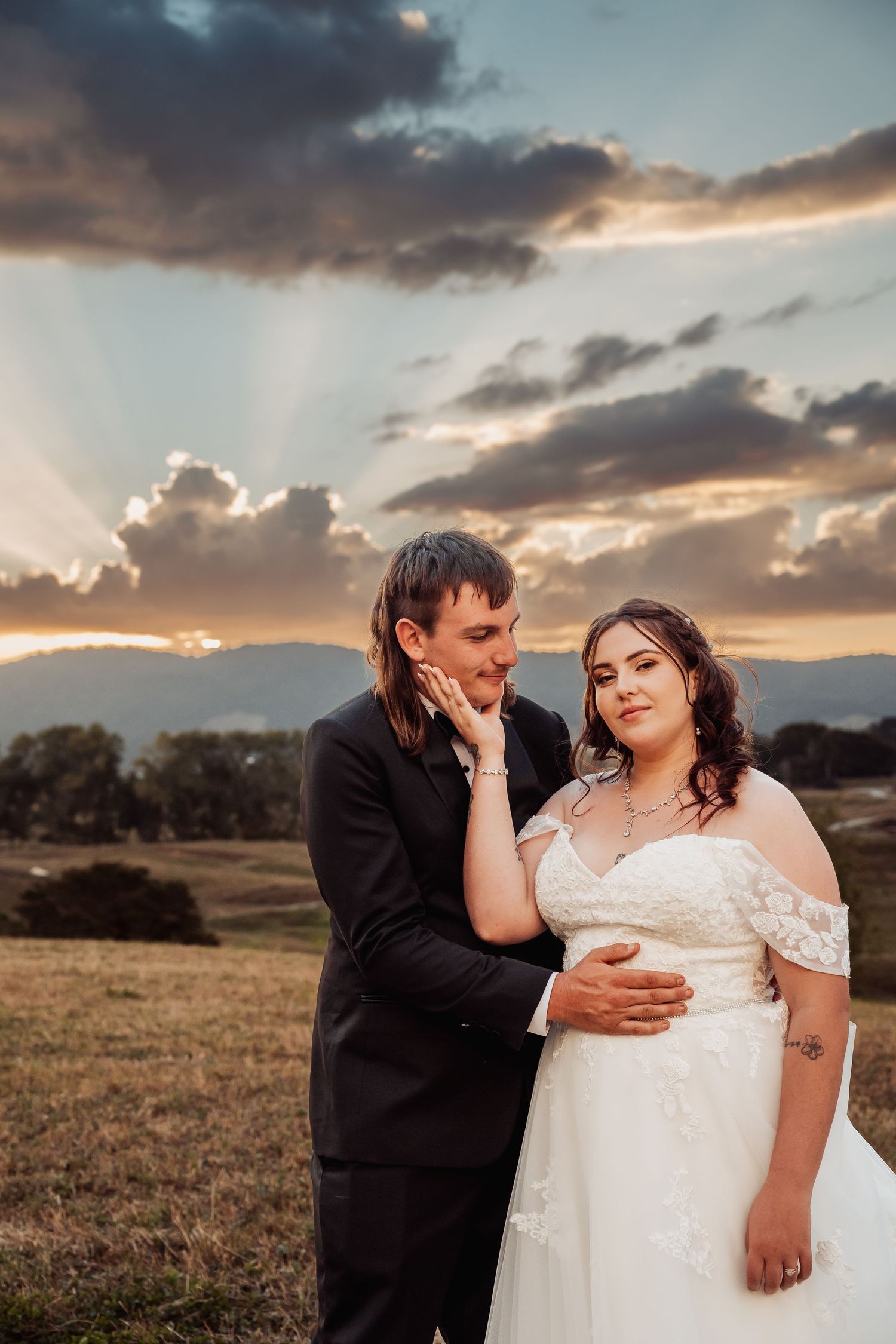 Groom tenderly holds bride's face as they pose together in a field under dramatic golden sunset rays.