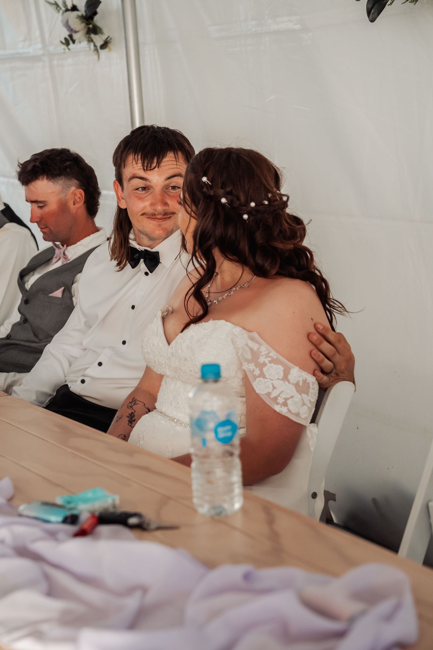 Newlywed groom smiling and embracing bride in lace gown at wedding reception table.