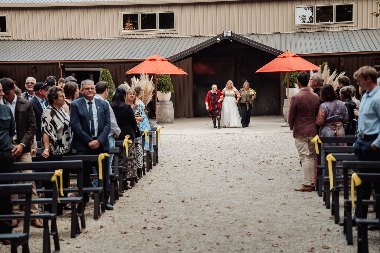 Wedding ceremony in rustic barn venue with guests seated in wooden pews along sandy aisle with orange umbrellas.