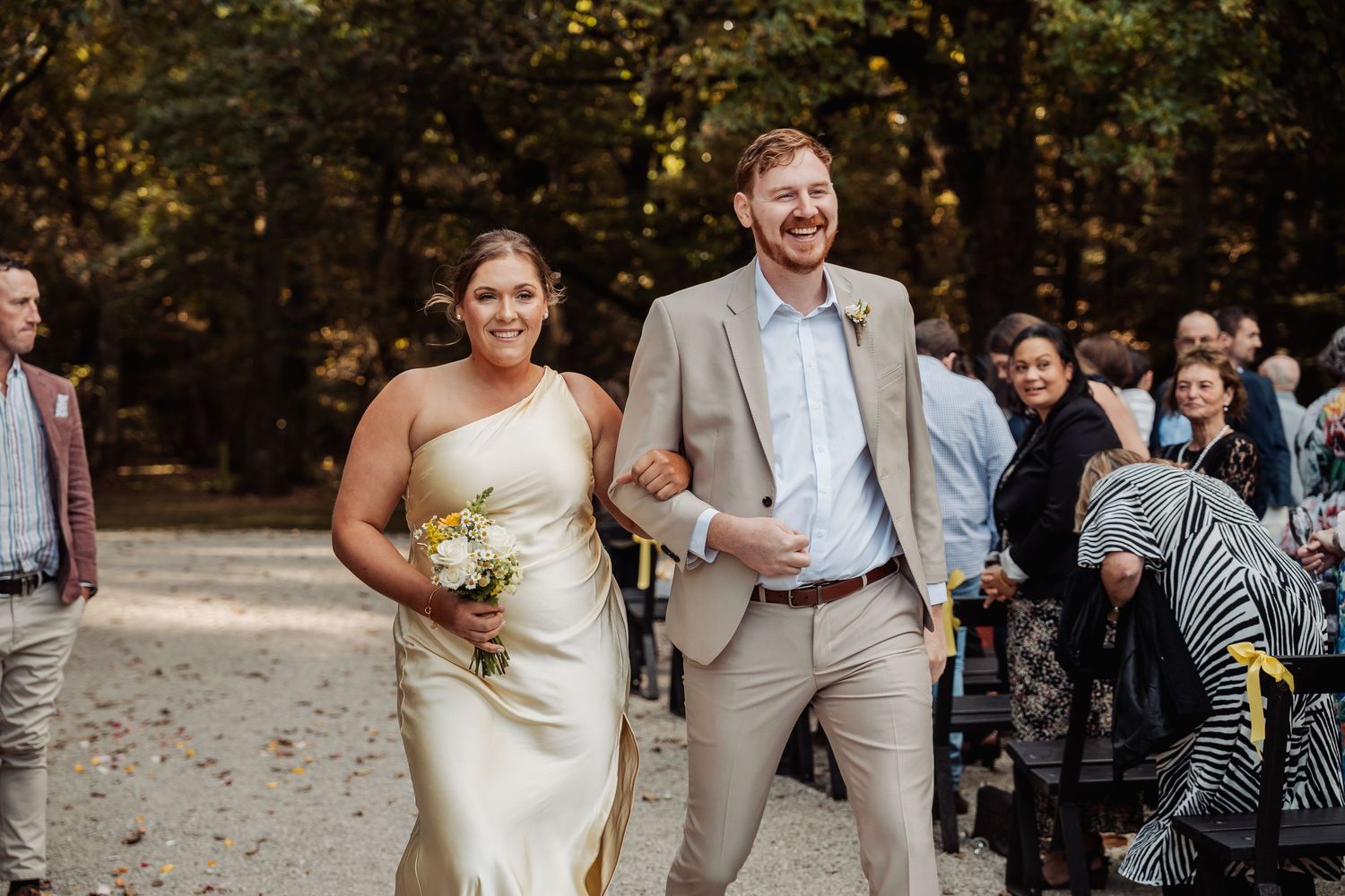 Bride in gold dress and groom in tan suit walking happily down outdoor aisle.