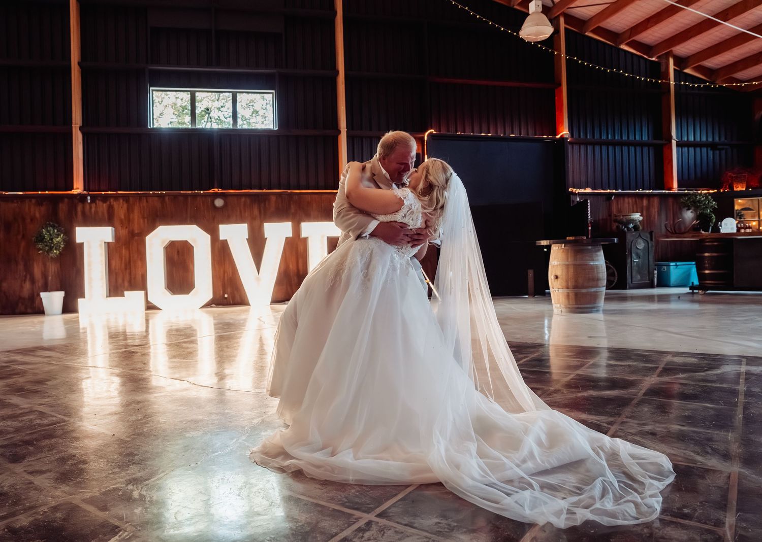 Bride and groom share first dance in rustic barn venue with illuminated LOVE letters backdrop.