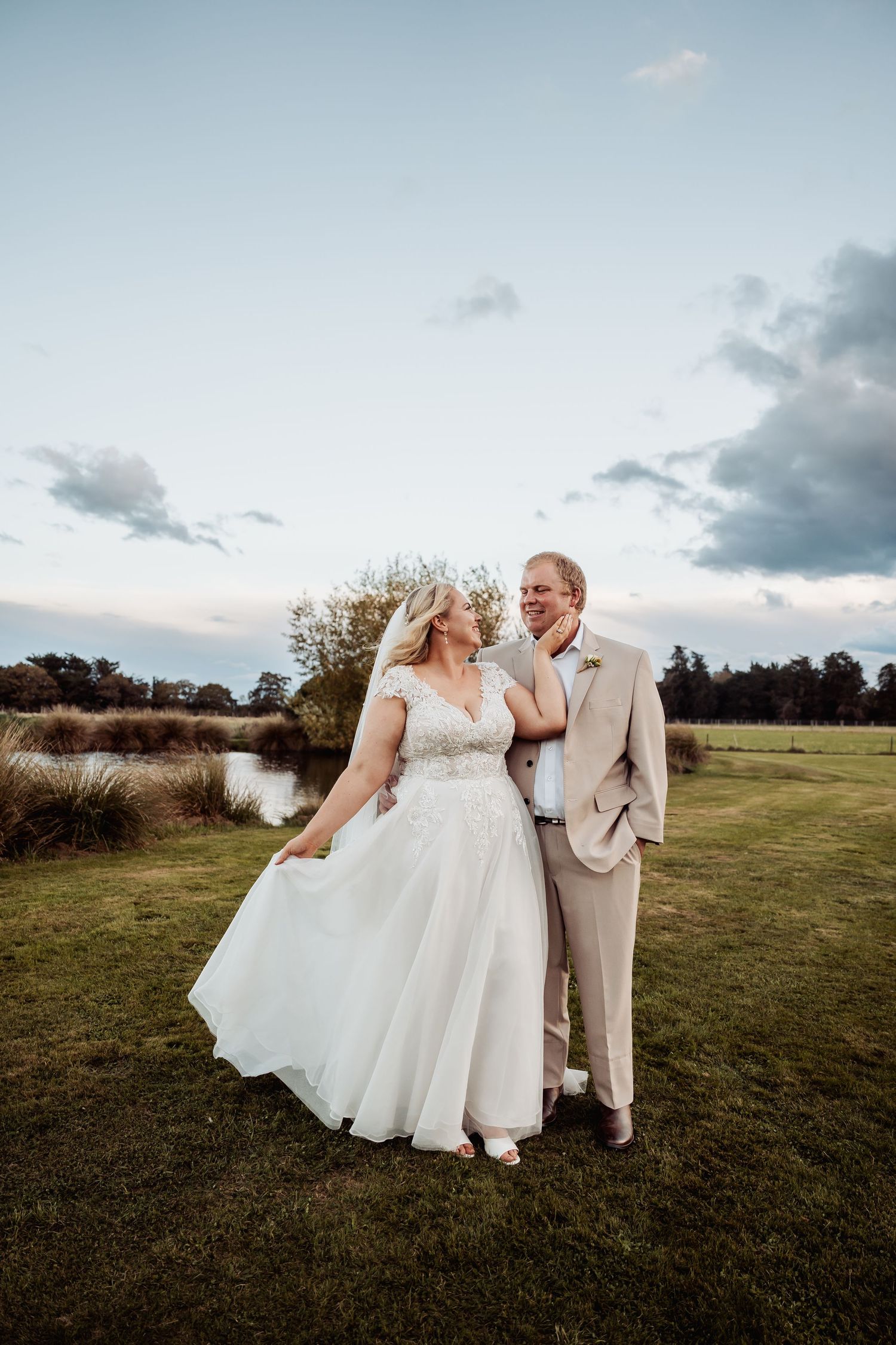 Bride and groom share romantic moment on green lawn at sunset near a peaceful pond.