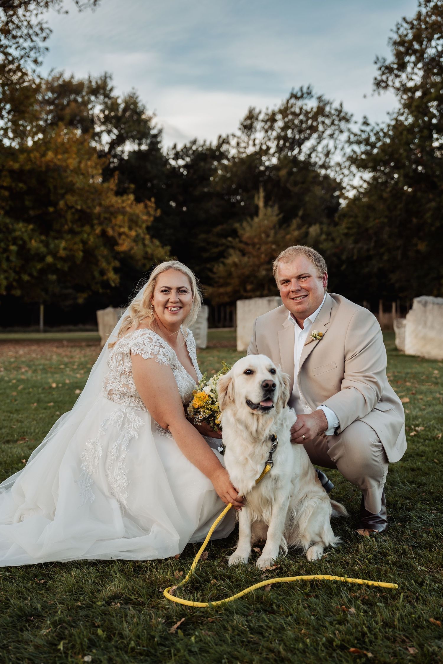 Bride and groom posing with white golden retriever dog on grass at outdoor wedding.