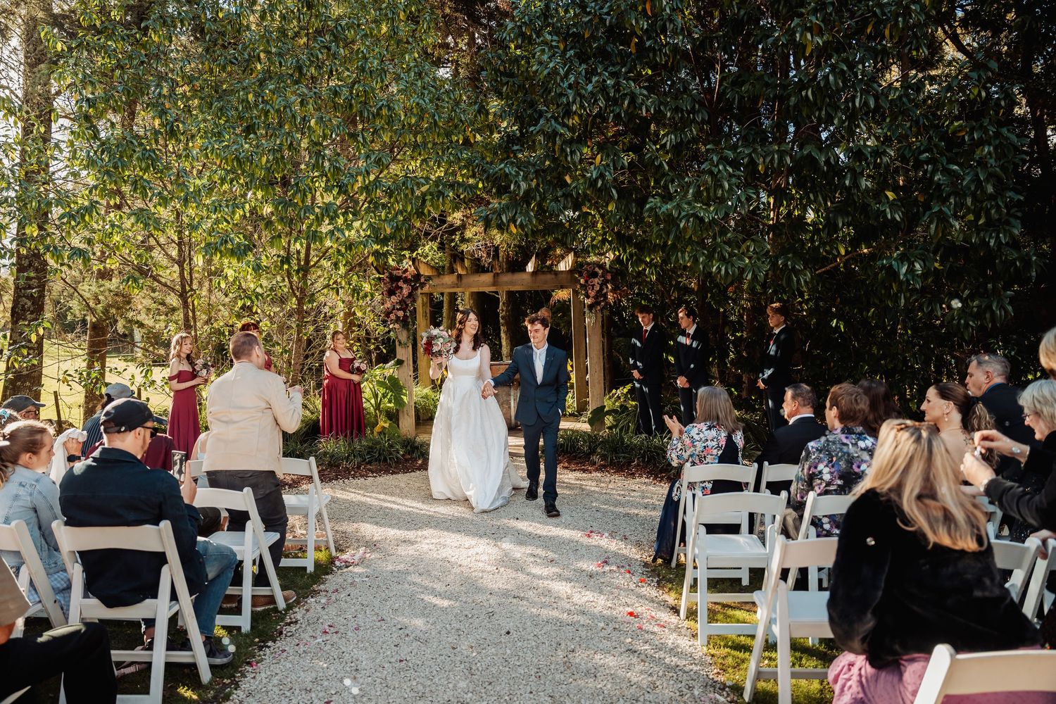 Bride and groom walking down outdoor wedding aisle surrounded by seated guests.