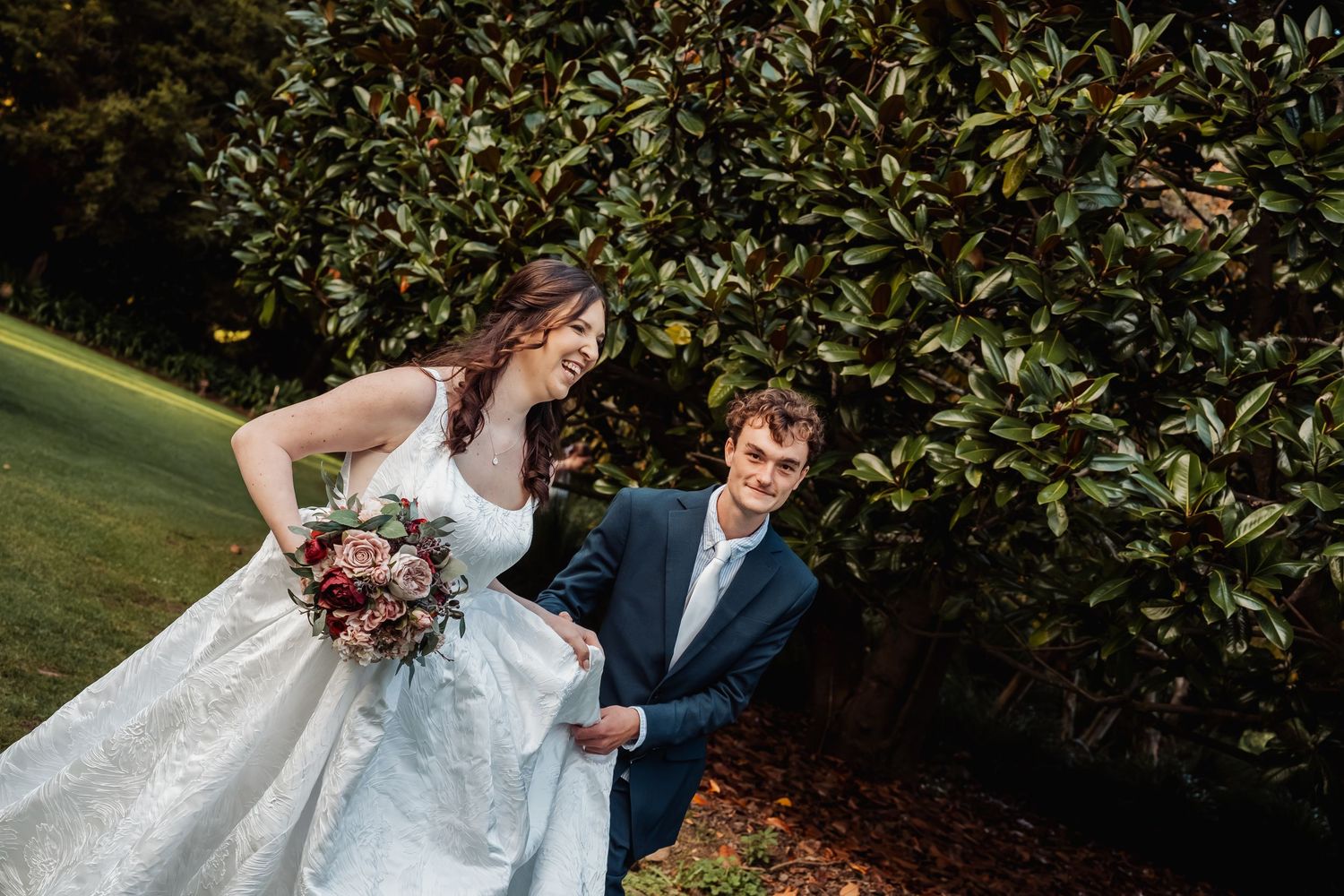 Bride in white gown laughing with groom in navy suit beside a large green hedge outdoors.
