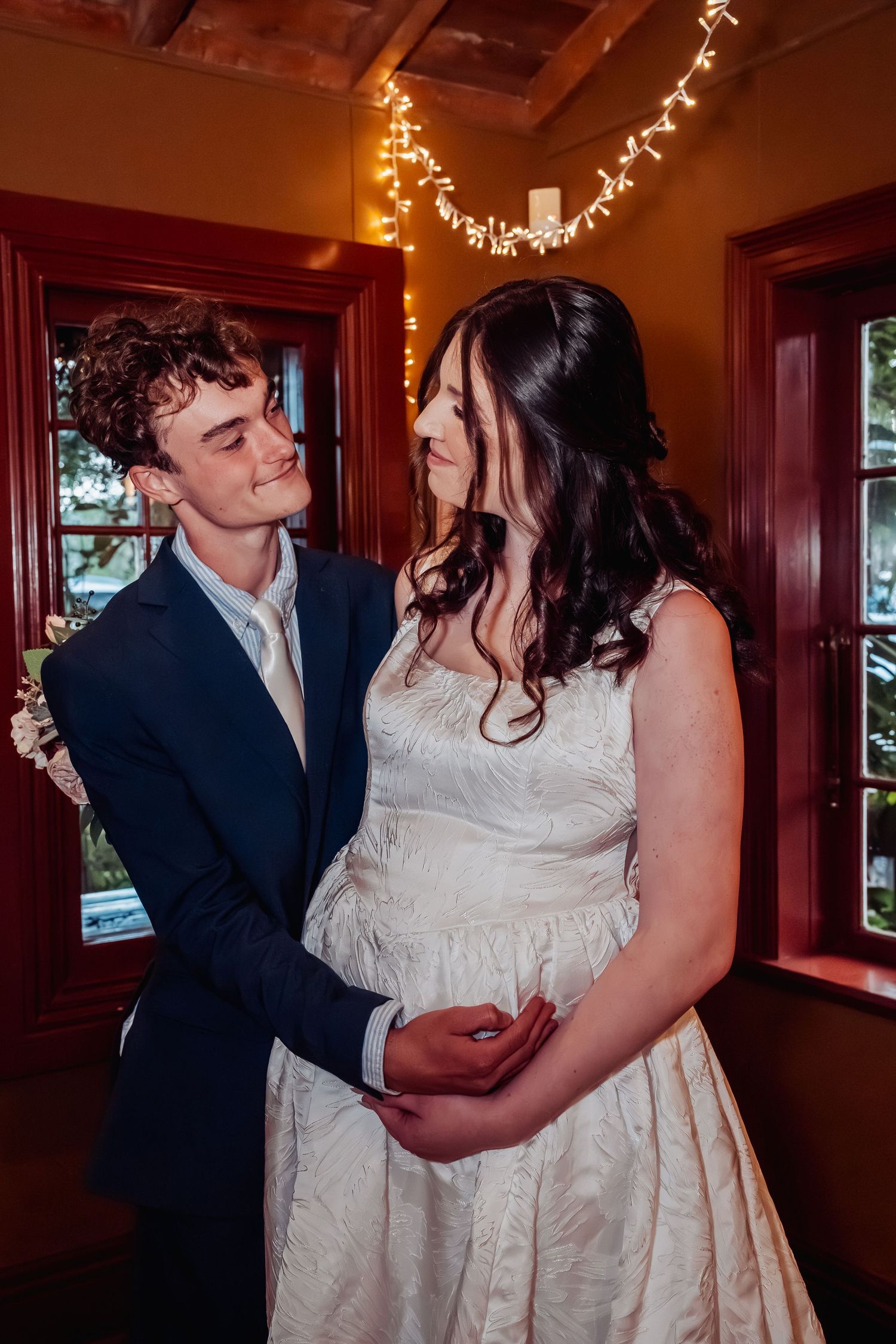 Newlyweds share intimate moment indoors with fairy lights glowing behind them.